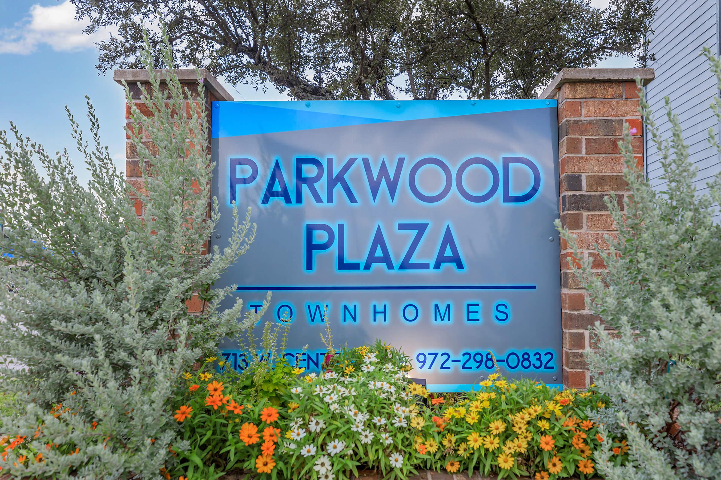 A prominent sign for Parkwood Plaza Townhomes, featuring blue and silver lettering on a large background. The sign includes contact information and is surrounded by green plants and colorful flowers, set against a clear sky.