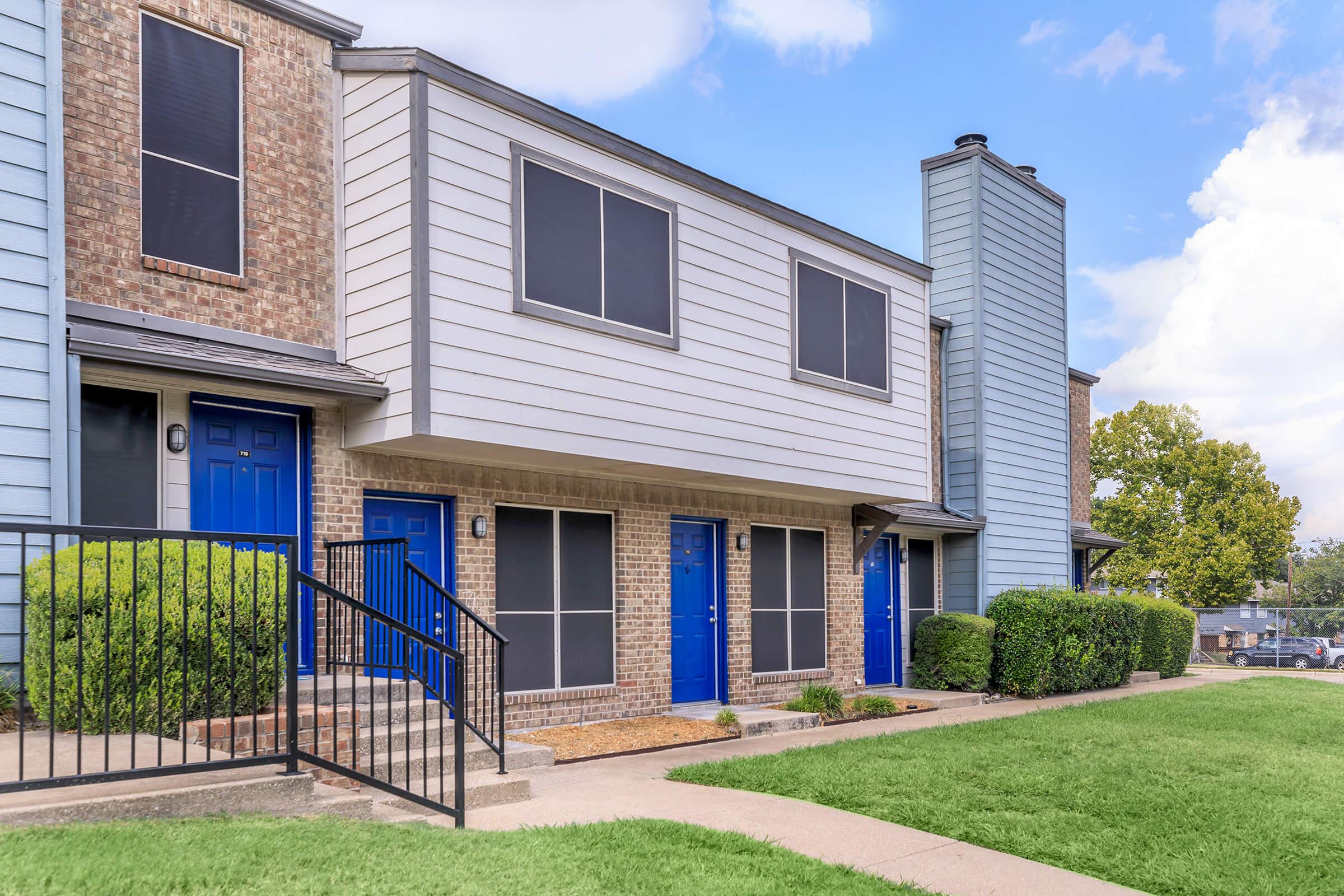 Row of modern townhouses featuring a mix of brick and siding exteriors. Each unit has distinctive blue doors and window shutters. Lush green grass surrounds the pathway leading to the entrance, with well-maintained shrubs adding to the curb appeal. Clear blue sky in the background.
