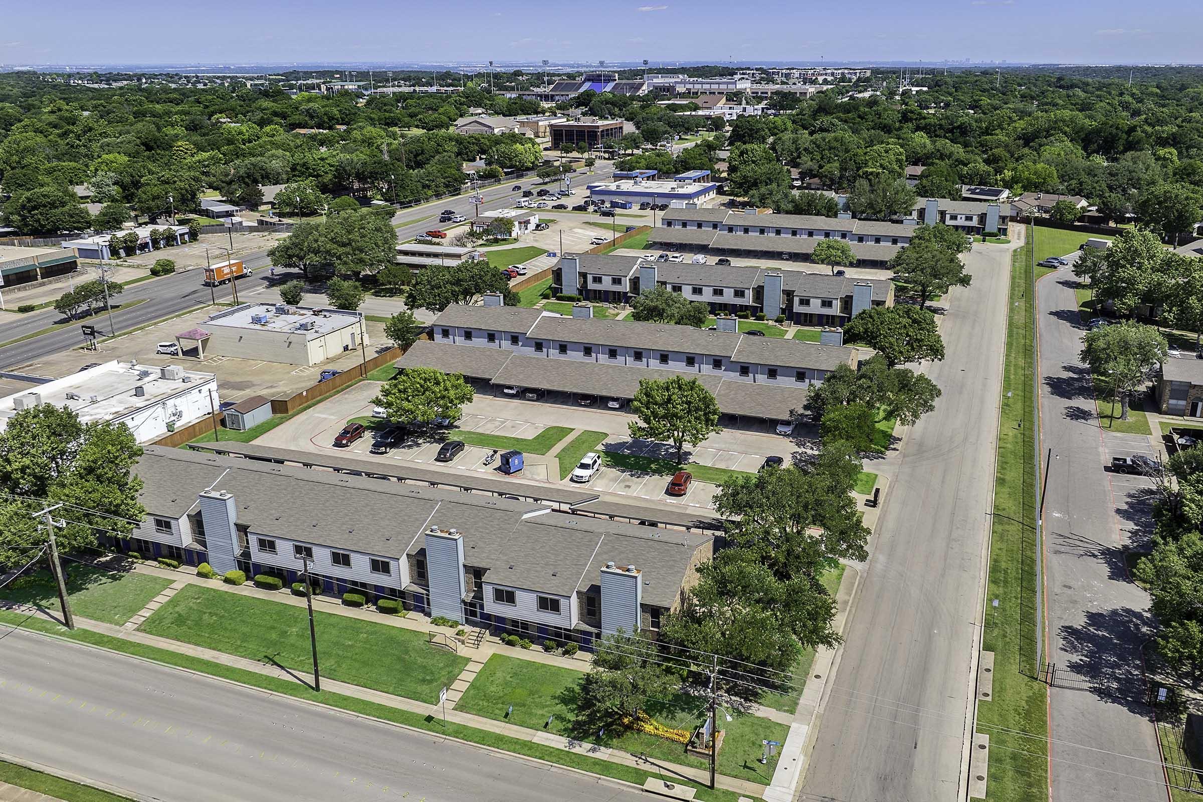 An aerial view of a suburban neighborhood featuring several low-rise apartment buildings with roofs, surrounded by green lawns and trees. The street runs parallel to the buildings, with parked cars visible. In the background, urban developments and a blue sky with scattered clouds are visible.