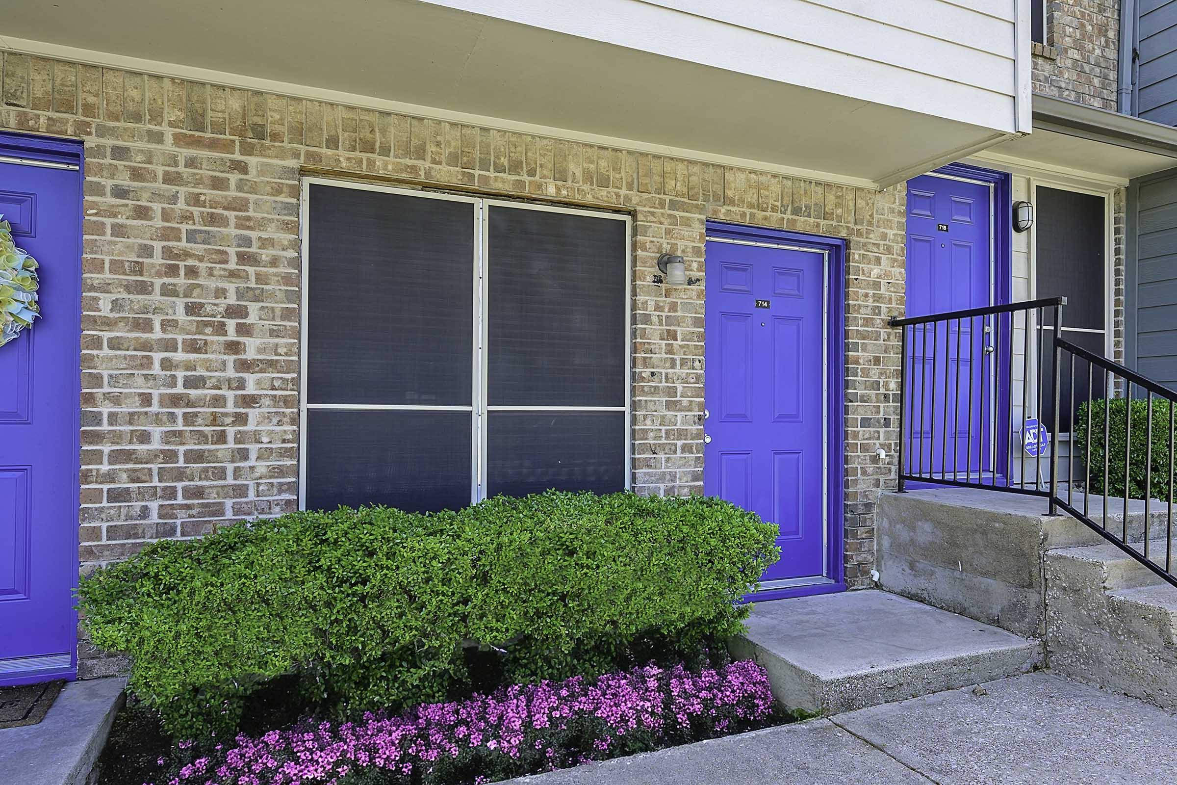 Exterior view of a brick apartment building featuring two bright blue doors. A screened window is positioned between the doors, with a neatly trimmed green hedge and pink flowers lining the walkway. A staircase with a black railing leads to the upper level, enhancing the entrance's appeal.