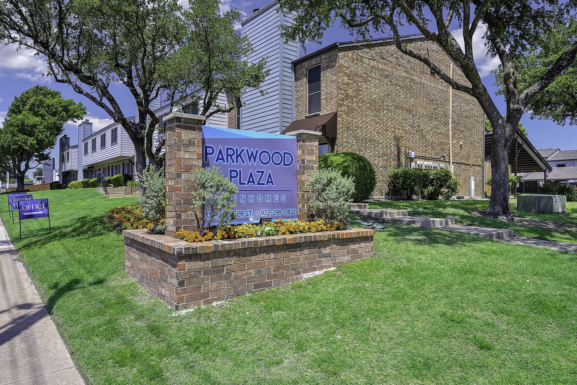 Sign for Parkwood Plaza surrounded by green grass and trees, with a brick structure and colorful flower beds. Nearby buildings in the background indicate a residential area. Clear blue skies add to the pleasant atmosphere.