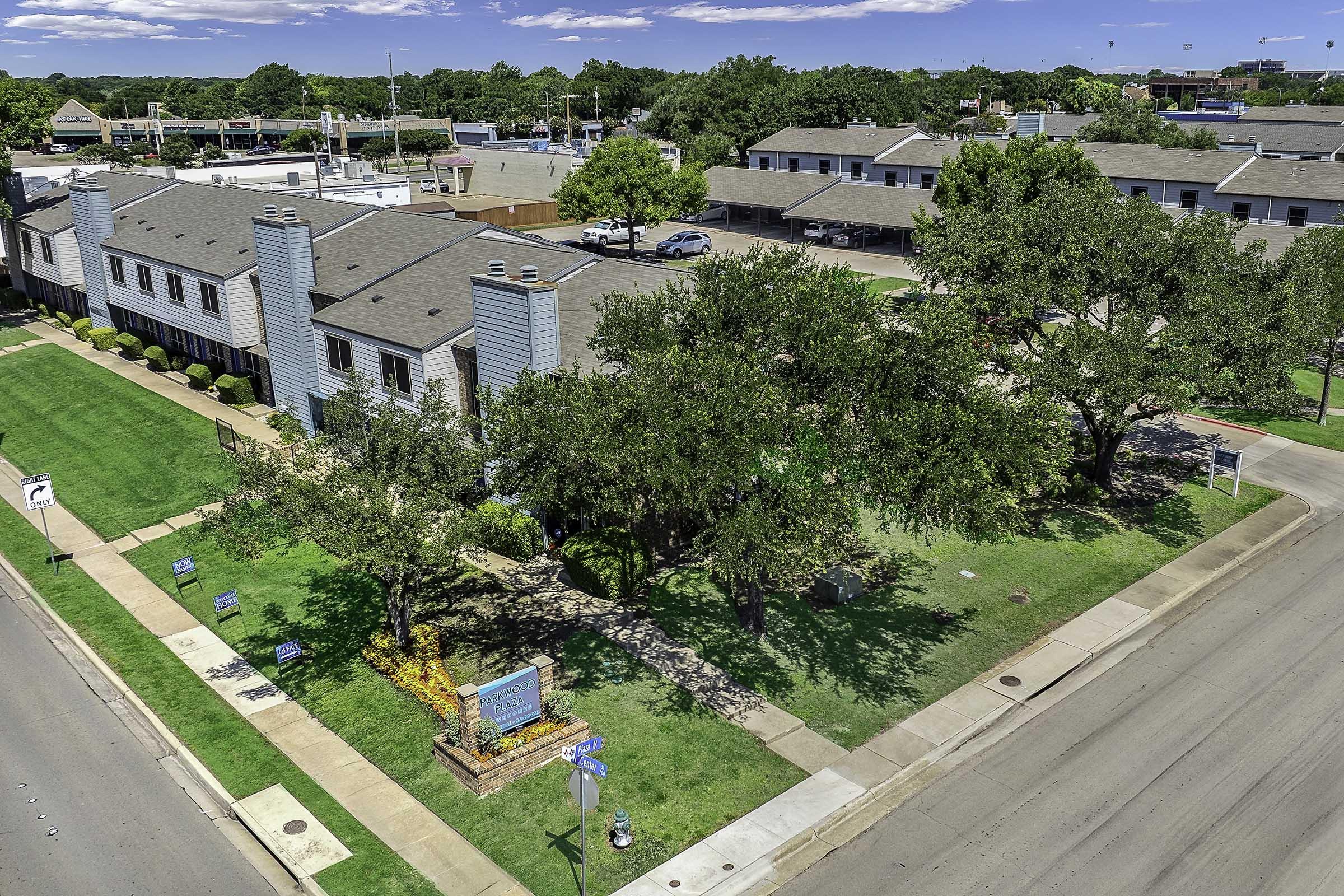 Aerial view of a multi-story building with light blue siding, surrounded by neatly trimmed lawns and trees. The area is well-maintained, with signage visible near the entrance. Nearby streets and buildings can be seen in the background under a clear blue sky.