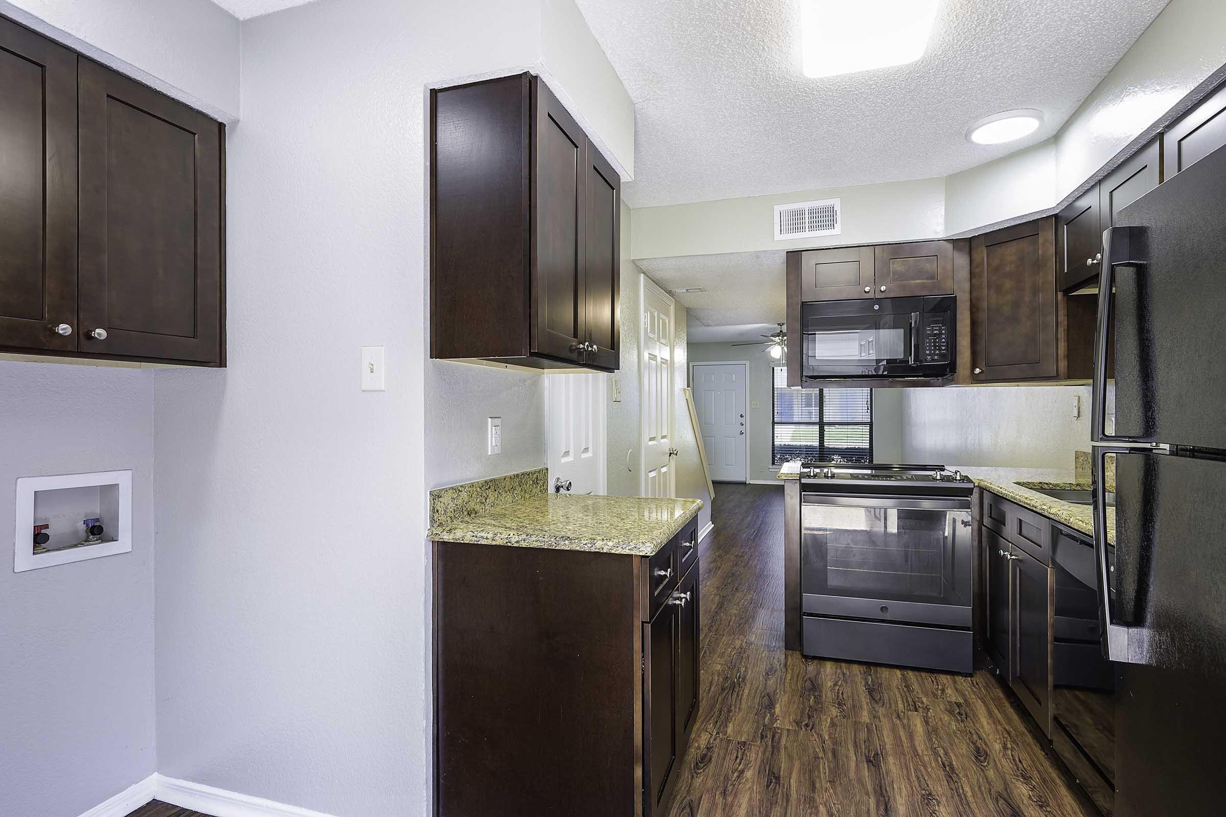 Modern kitchen featuring dark wooden cabinets, granite countertops, stainless steel appliances including a microwave, stove, and refrigerator. The open layout leads to a well-lit area with laminate flooring and a doorway visible in the background.