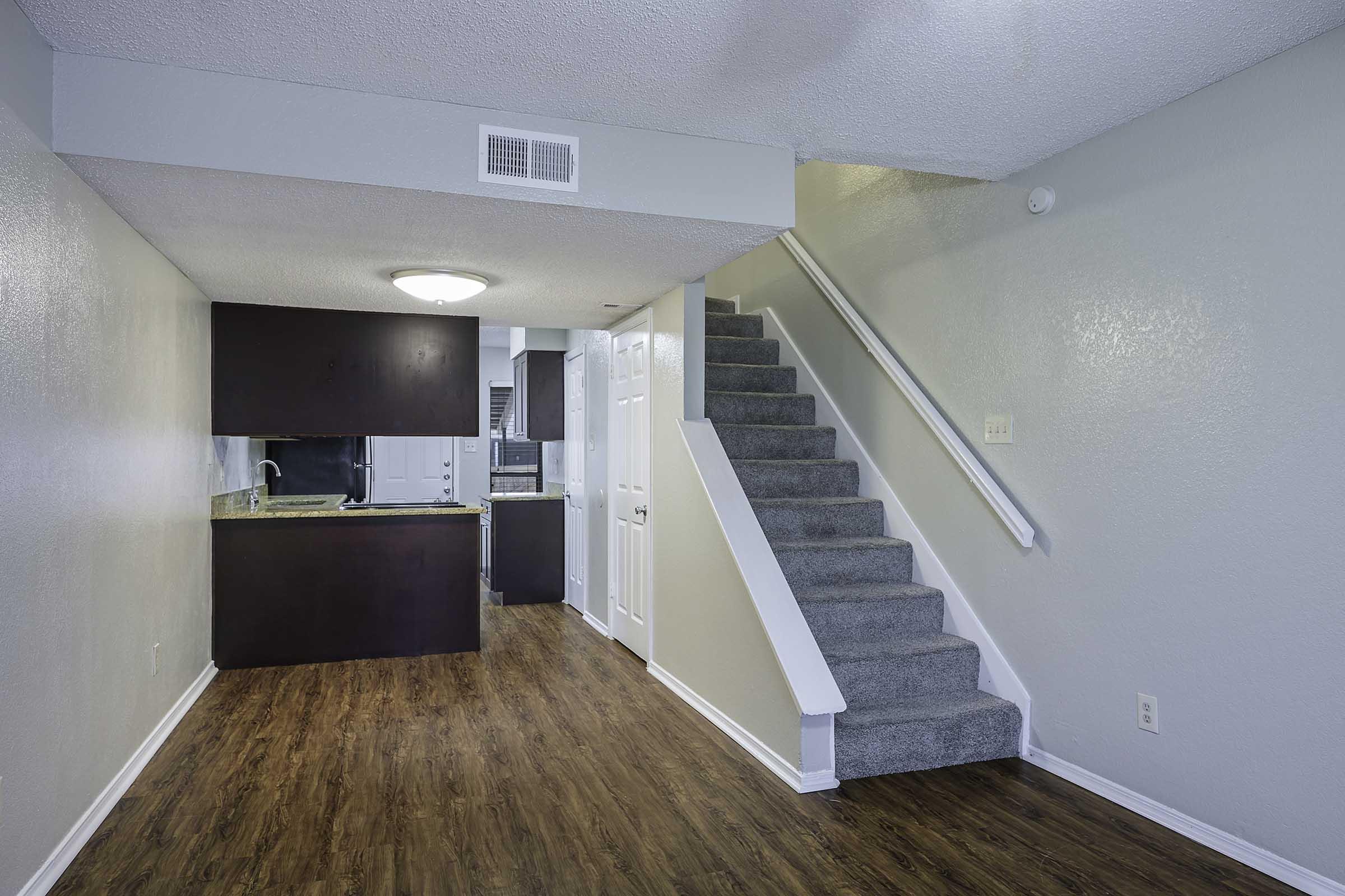 Interior view of a modern apartment. The room features a staircase leading to the upper level, hardwood flooring, and a kitchen area with dark cabinets. The walls are painted in neutral tones, and there is a ceiling light fixture illuminating the space.
