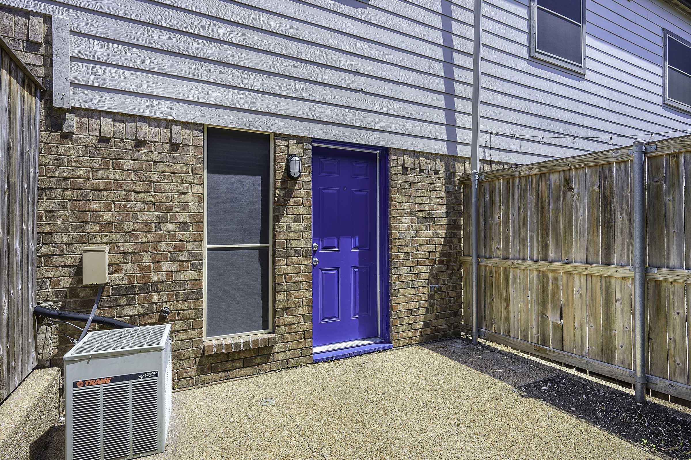 A blue door in a brick wall, surrounded by a wooden fence. An air conditioning unit is positioned next to the door, and the ground is covered with gravel. There are windows visible on the side of the building, and the setting appears to be an outdoor area of a residential property.