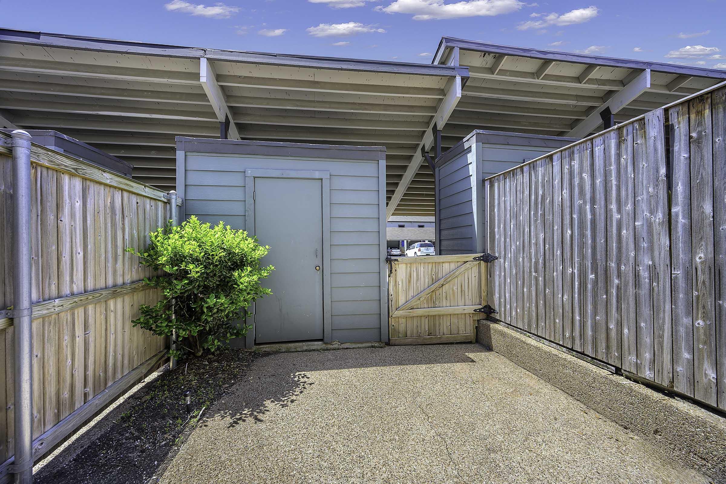 A narrow pathway between two wooden fences leads to a light blue door. On one side, there's a small shrub, and on the other, a gate. The area is characterized by a concrete surface under a clear blue sky with a few clouds. The setting appears to be a private outdoor space.