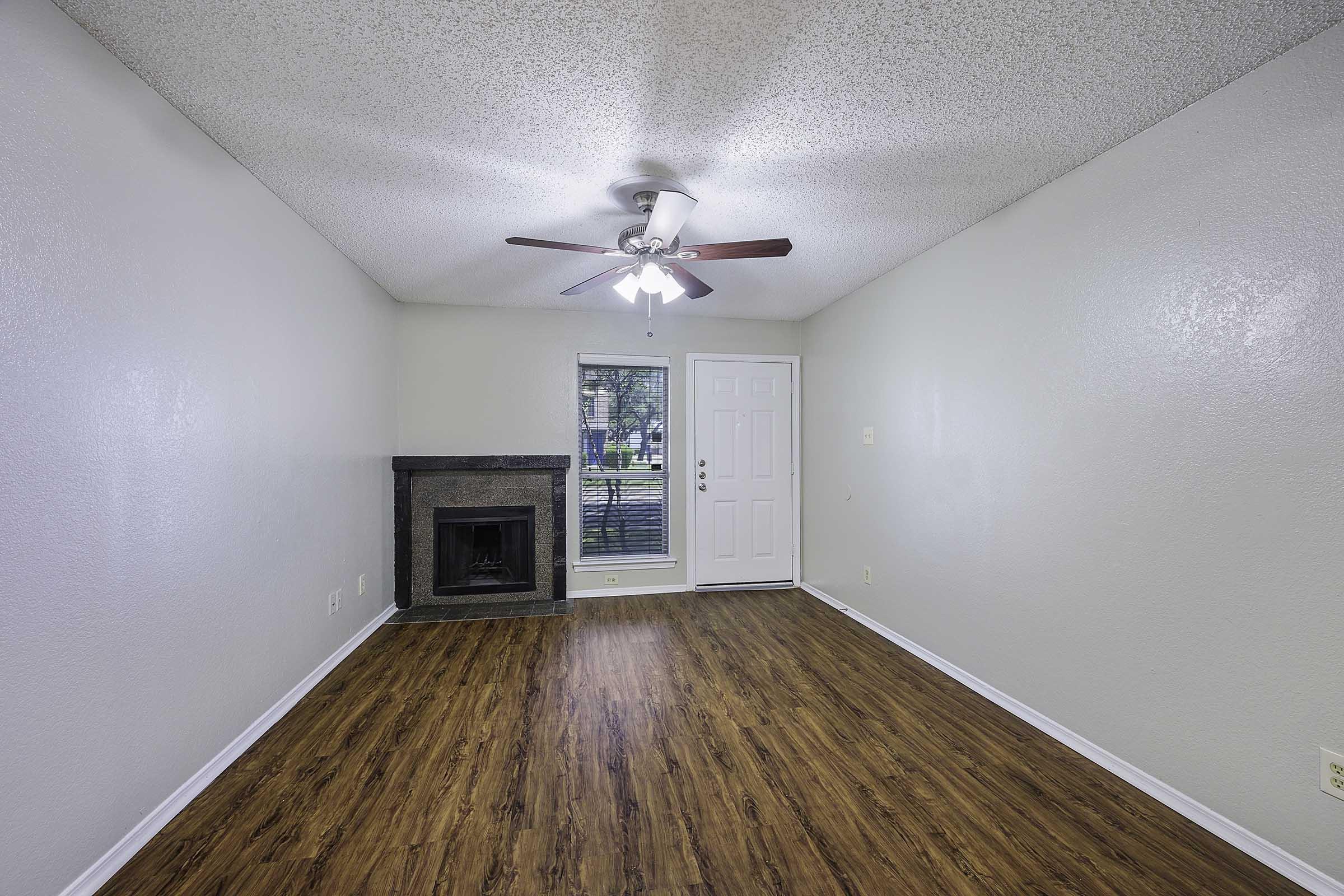 An empty living room showcasing a ceiling fan, light-colored walls, and a wooden floor. There is a fireplace on the left side and a front door with a window. Natural light filters in through the window, illuminating the space.