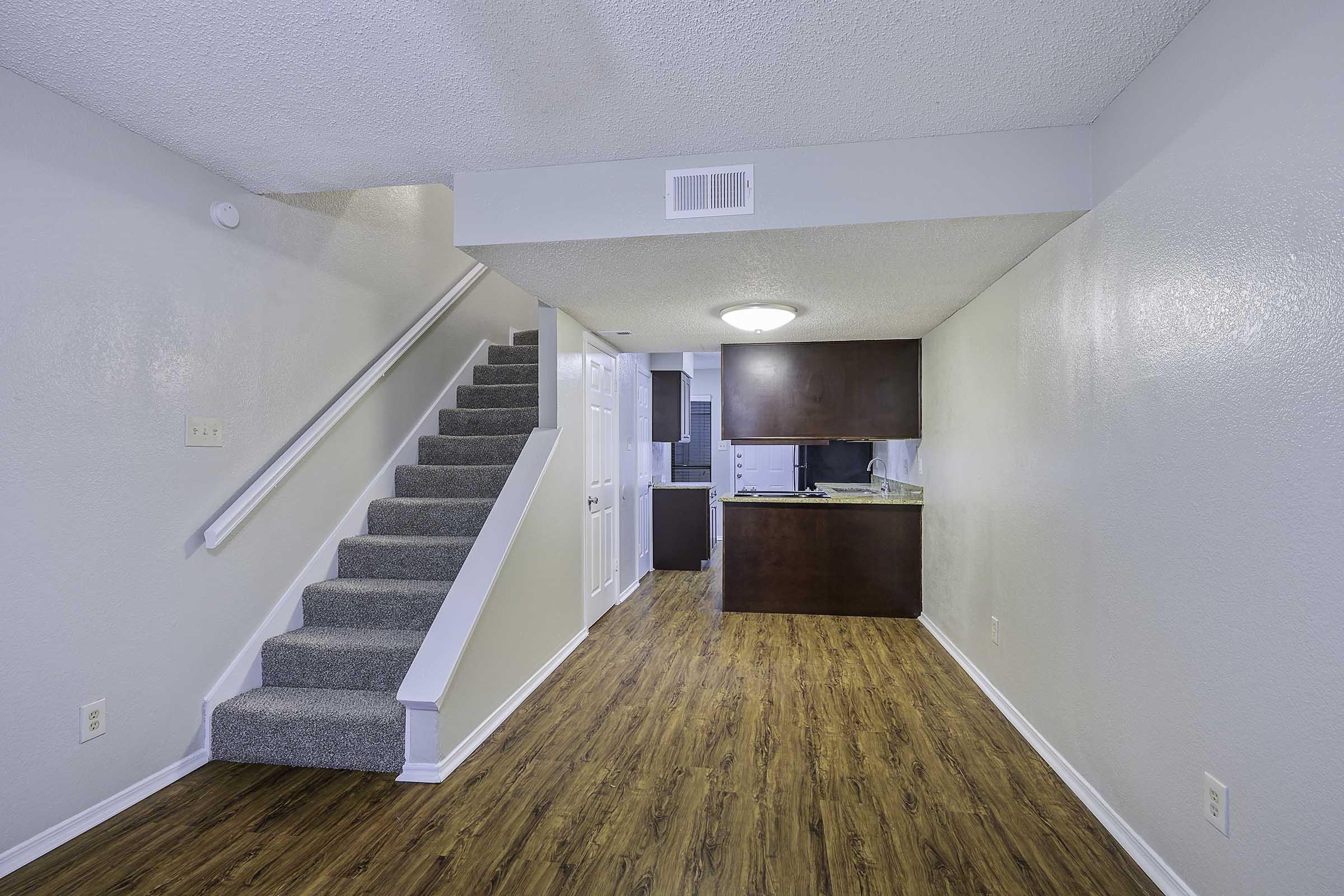 Interior view of a modern apartment featuring a staircase leading to the upper floor. The room has light-colored walls and a wood-like floor, with a kitchen area visible in the background. Bright lighting enhances the spacious feel of the living space.