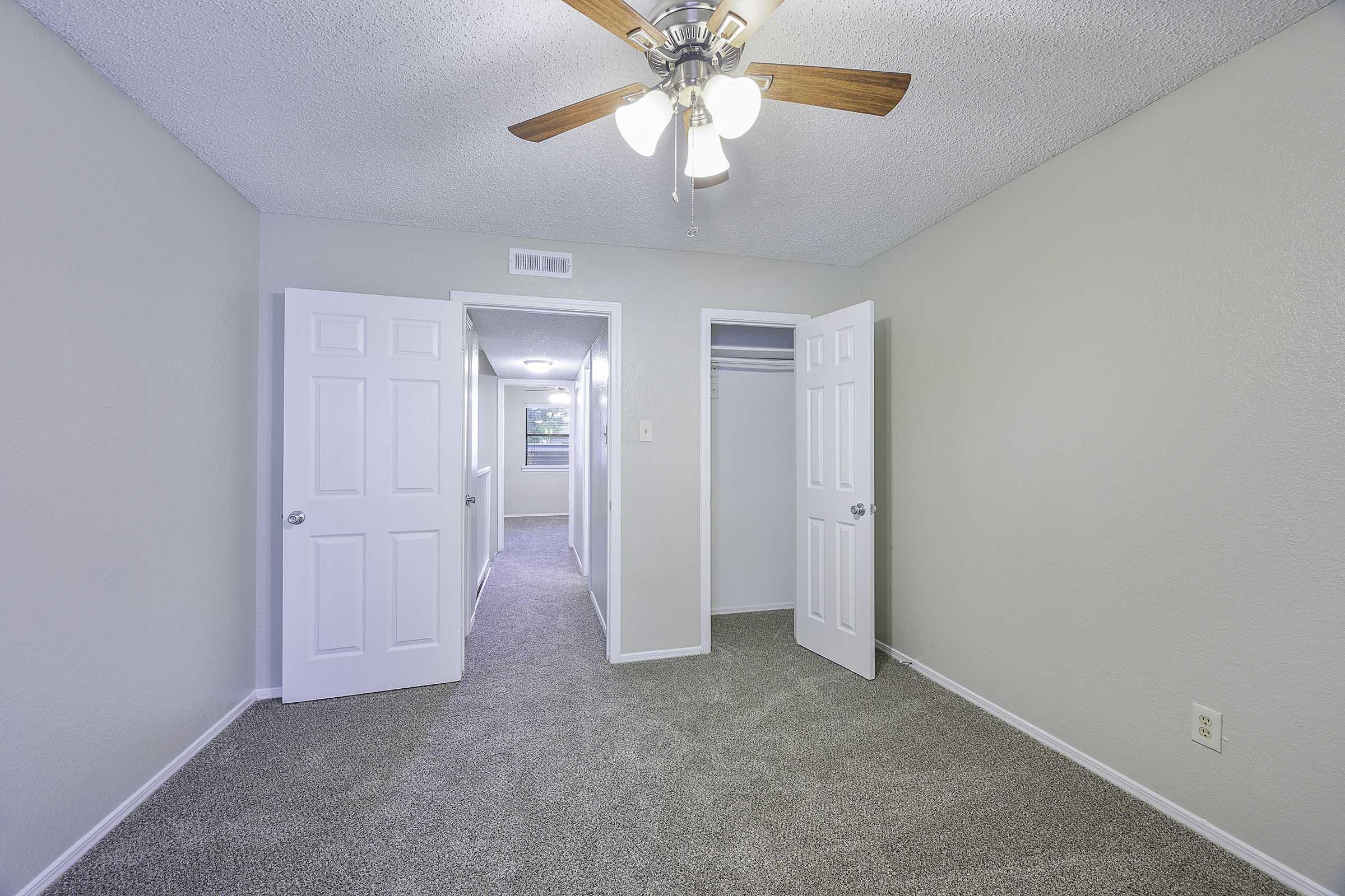A clean, well-lit bedroom featuring light gray walls and plush carpet. A ceiling fan with wooden blades is mounted in the center. The room has two open doors leading to a hallway and a closet, creating an inviting space suitable for relaxing or sleeping.
