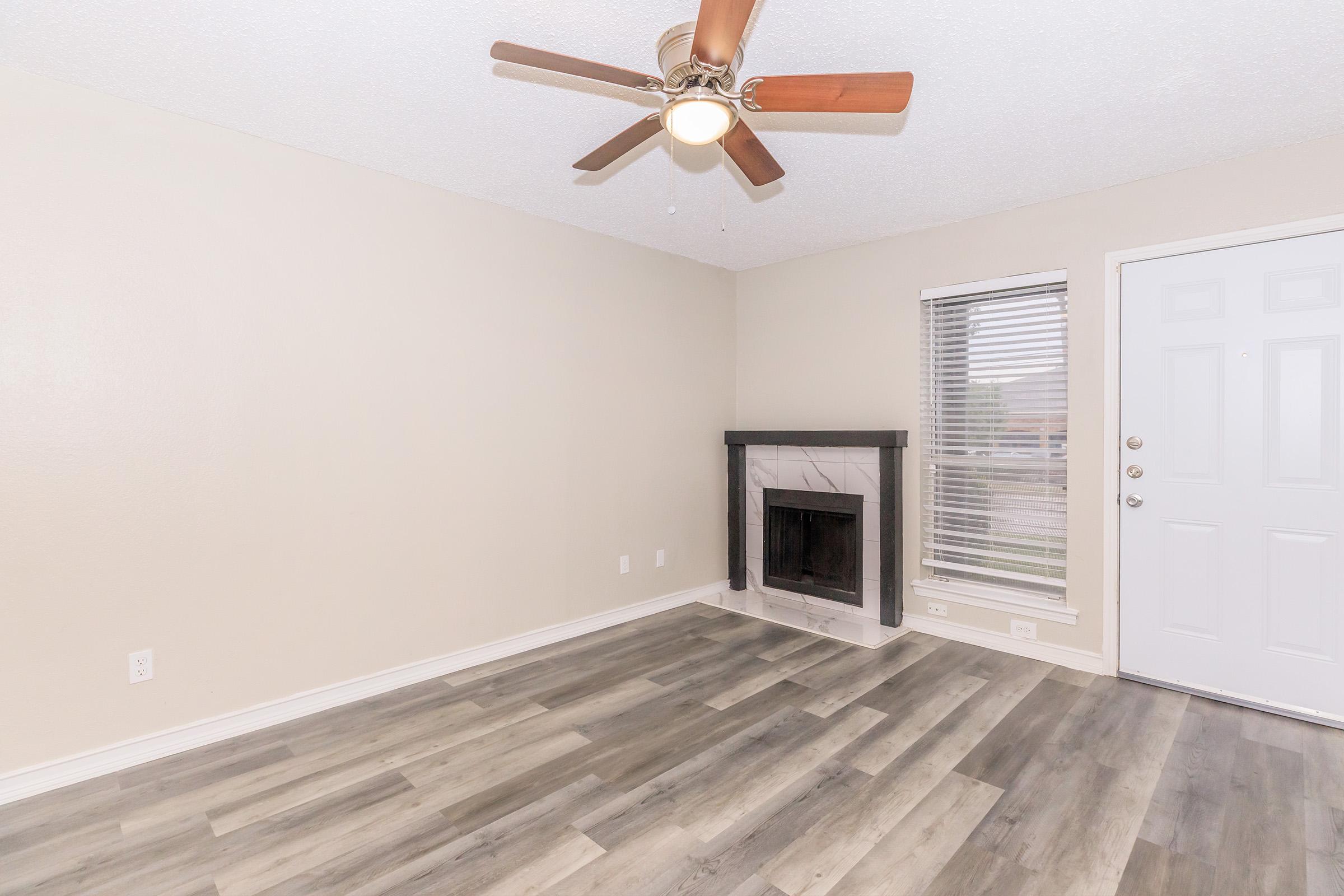 A spacious living room featuring a ceiling fan, light wood-like flooring, a fireplace with a black mantle, and a large window with blinds. A front door is visible on the right side, and the walls are painted in a neutral color. The room is well-lit and designed for comfort.