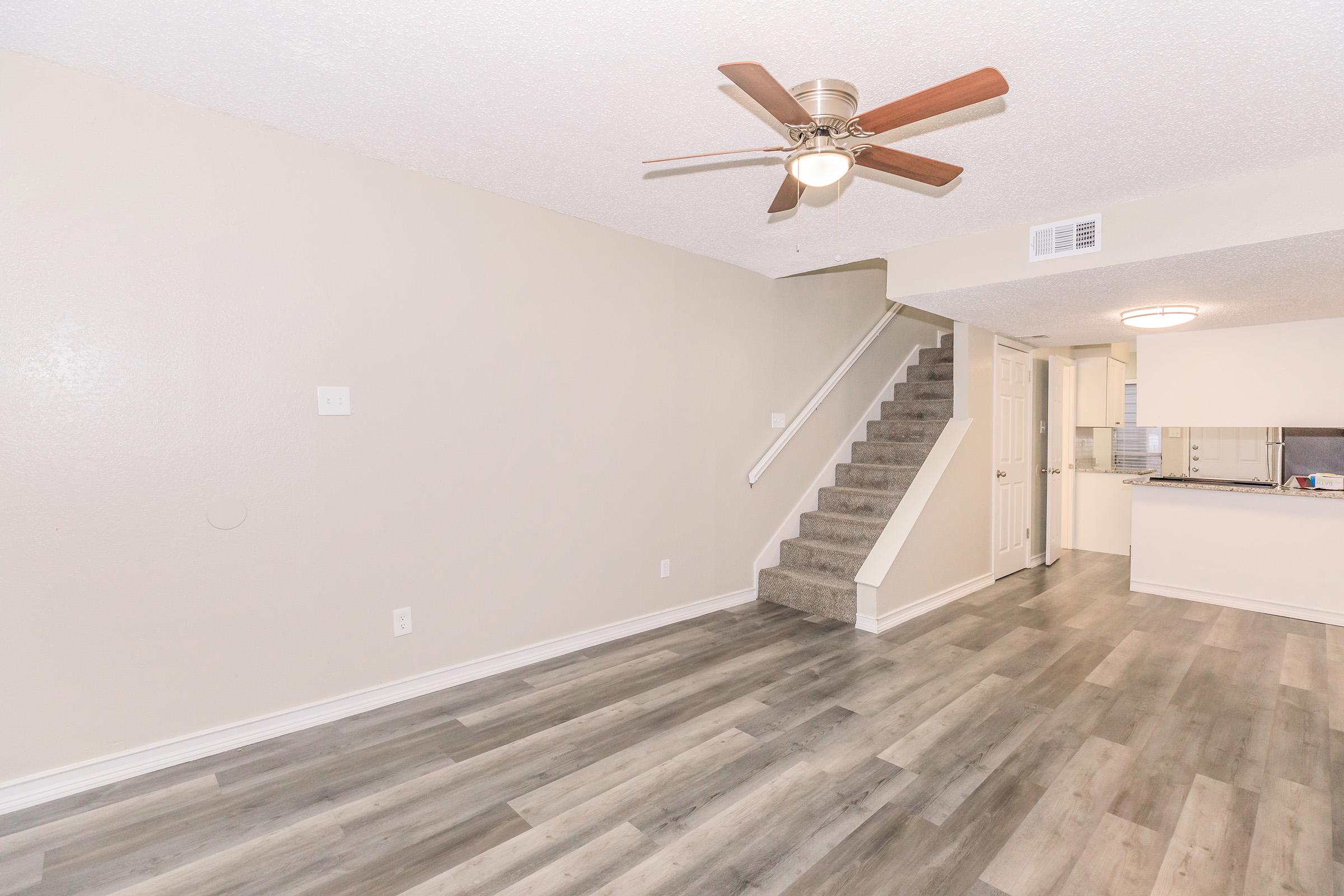 Spacious living area with light-colored walls and a ceiling fan. Features hardwood-style flooring and a staircase leading to an upper level. In the background, a glimpse of an open kitchen area with modern fixtures can be seen.