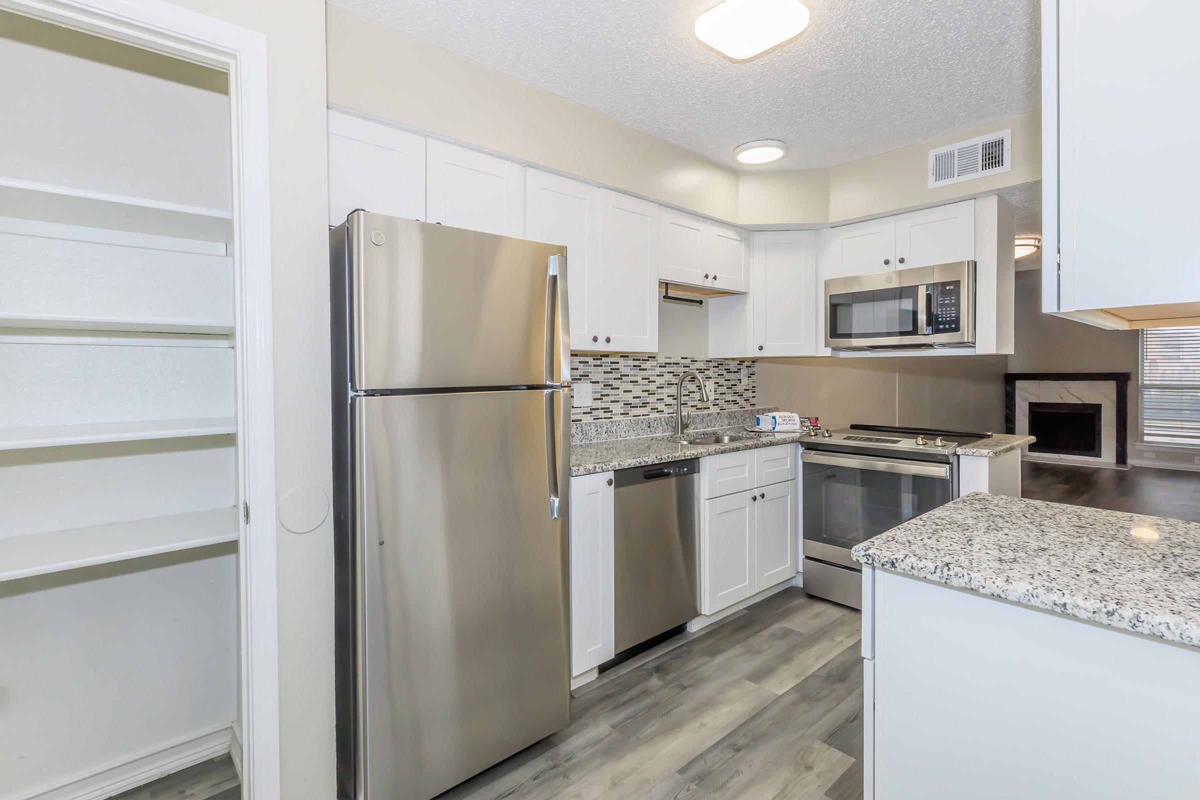 Modern kitchen featuring stainless steel appliances, white cabinetry, and a mosaic tile backsplash. The space includes a refrigerator, microwave, and oven, along with a spacious pantry on the left. Natural light fills the room, highlighting the granite countertops and gray laminate flooring.