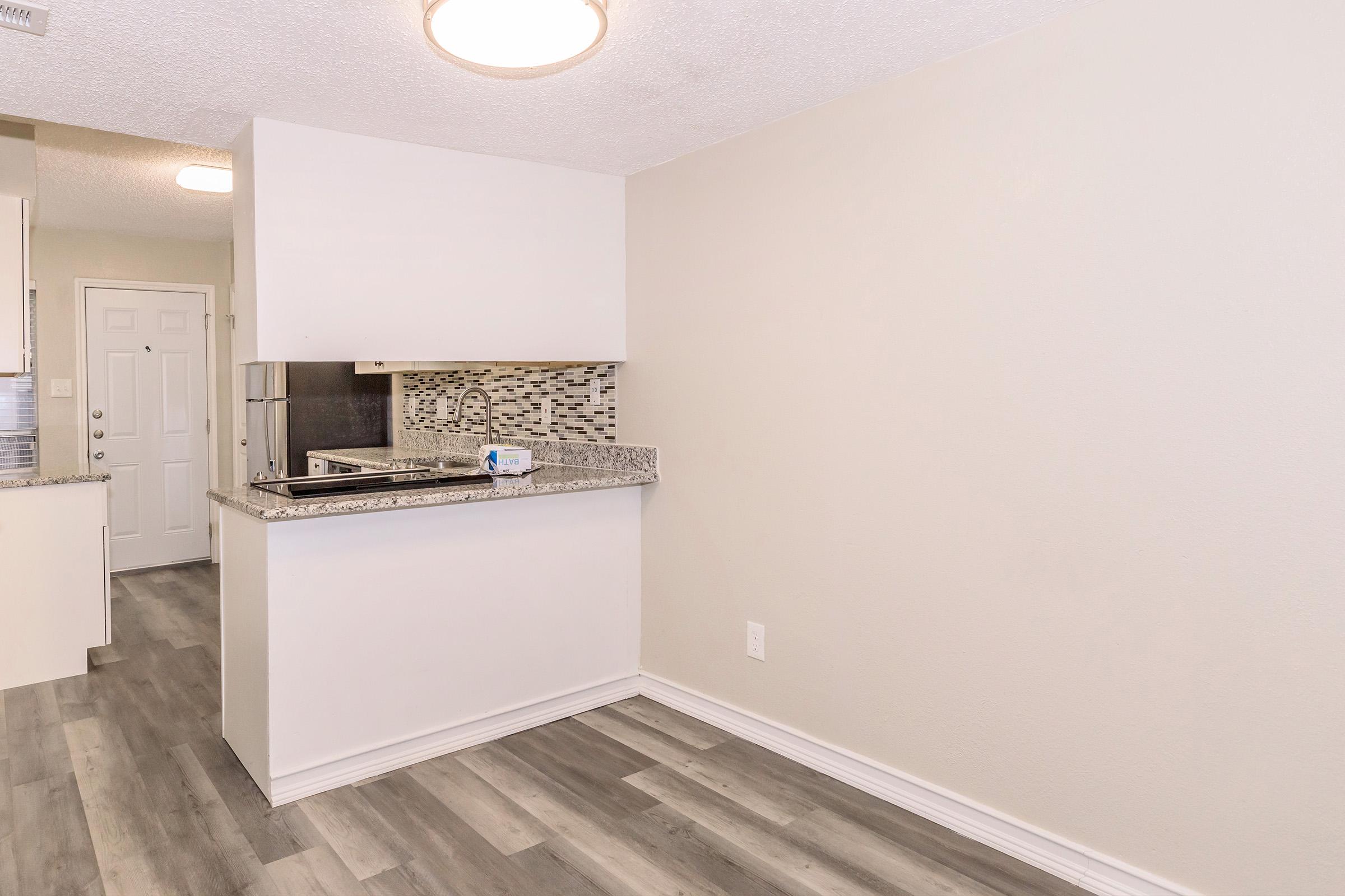 A modern kitchen space featuring light-colored walls, a granite countertop, and a mosaic backsplash. The flooring is a sleek wood-like laminate. There is an entry door visible, and the layout offers an open feel with ample natural light.