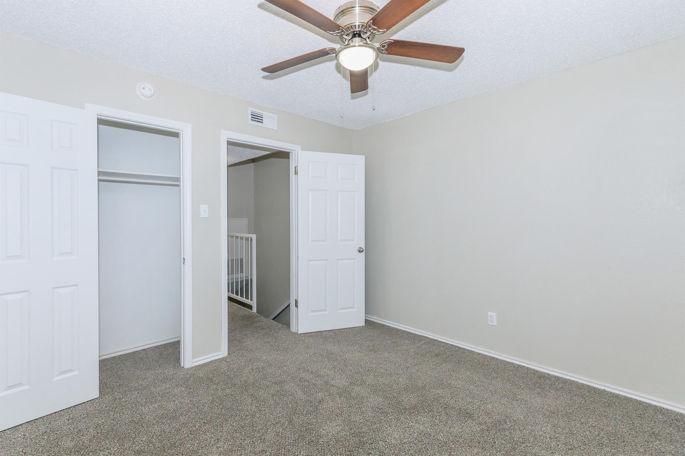 A spacious, empty bedroom featuring light beige walls, carpeted flooring, and a ceiling fan. There are two white double doors leading to a closet, and an open doorway visible in the background. Natural light is present, creating a bright and inviting atmosphere.