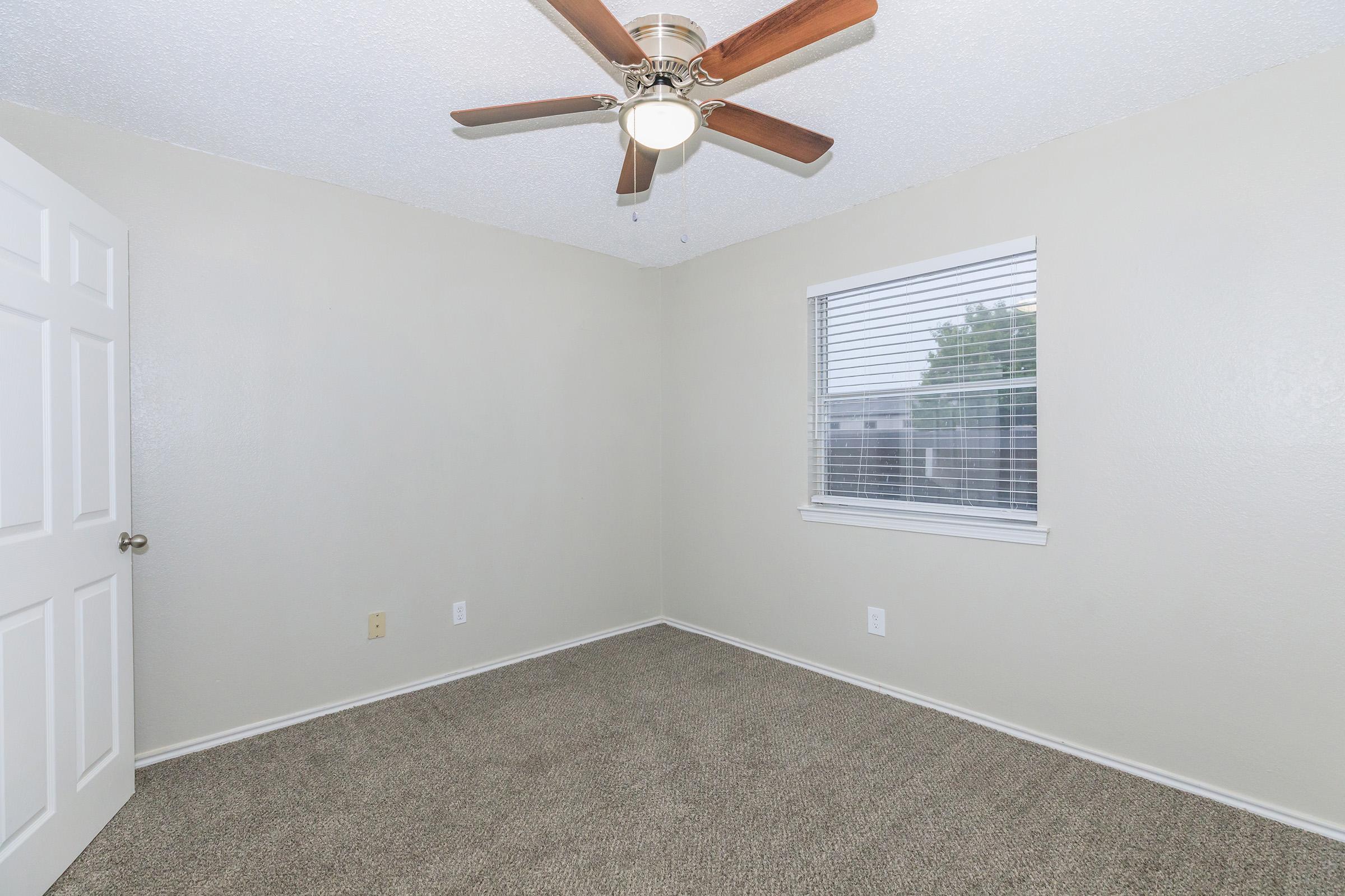 A small, empty room with light gray walls and beige carpet. The room features a ceiling fan with wooden blades, a window with white blinds, and a white door. Natural light enters through the window, illuminating the space.