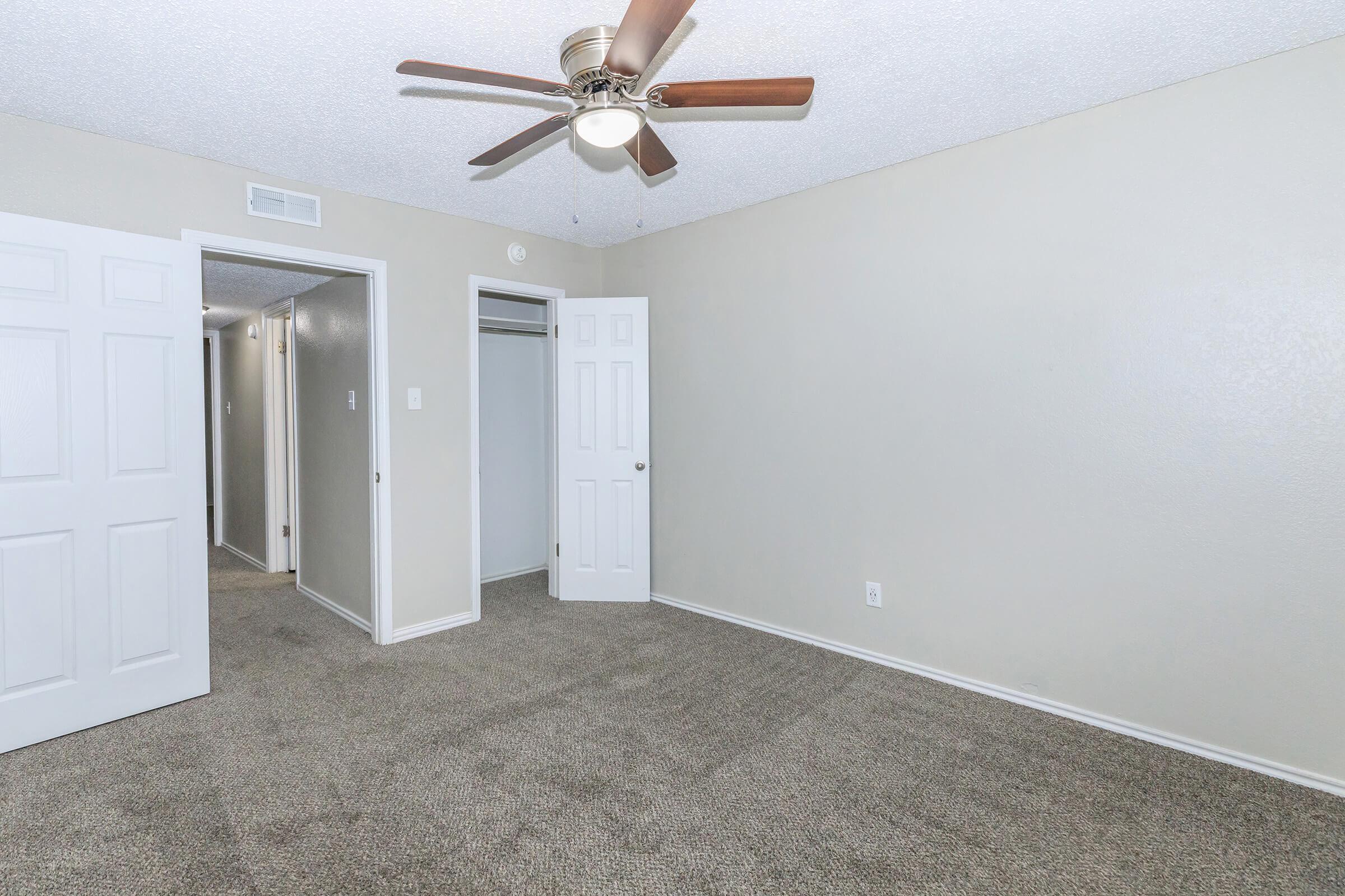 A spacious, carpeted bedroom featuring a ceiling fan, with light beige walls. To the left, there are two doors leading to closets and a hallway. The room is well-lit and has a clean, neutral aesthetic, making it suitable for various decor styles.