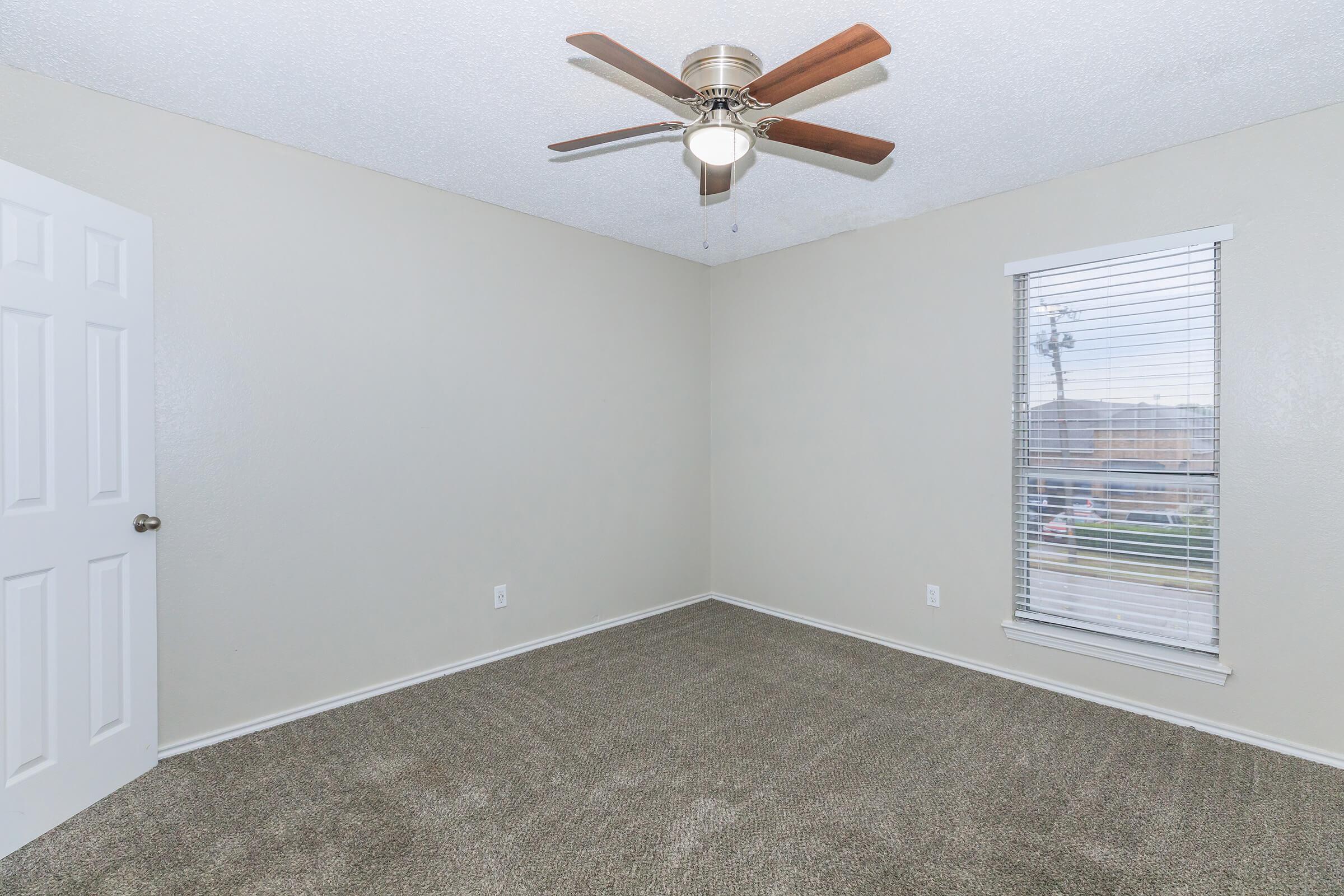 A clean, empty room with light beige walls and carpet. There is a ceiling fan with wooden blades, and a window with horizontal blinds allows natural light to enter. The room features a single door on the left and no furniture, creating a spacious feel. Ideal for customization or moving in furniture.