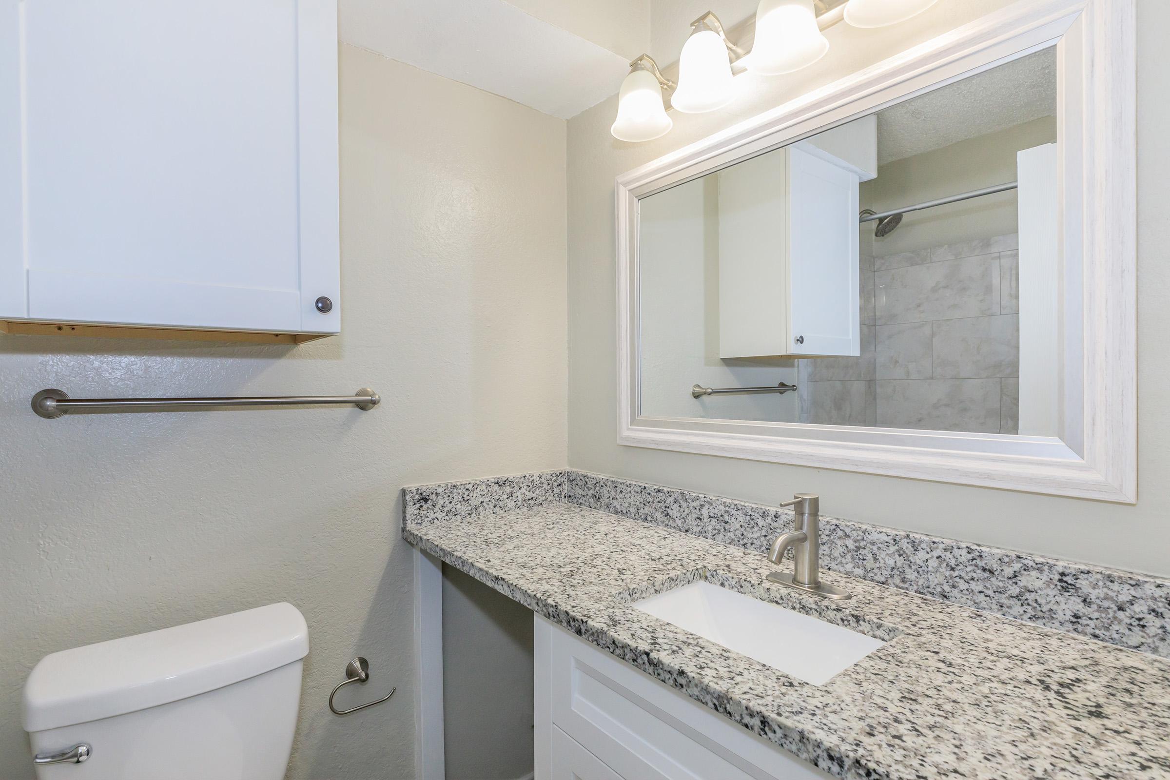 A modern bathroom featuring a white toilet, a sleek vanity with a white sink and a granite countertop. Above the sink, there is a large mirror framed in white, with three light fixtures illuminating the space. The walls are painted in a light color, and there is a towel bar mounted near the toilet.