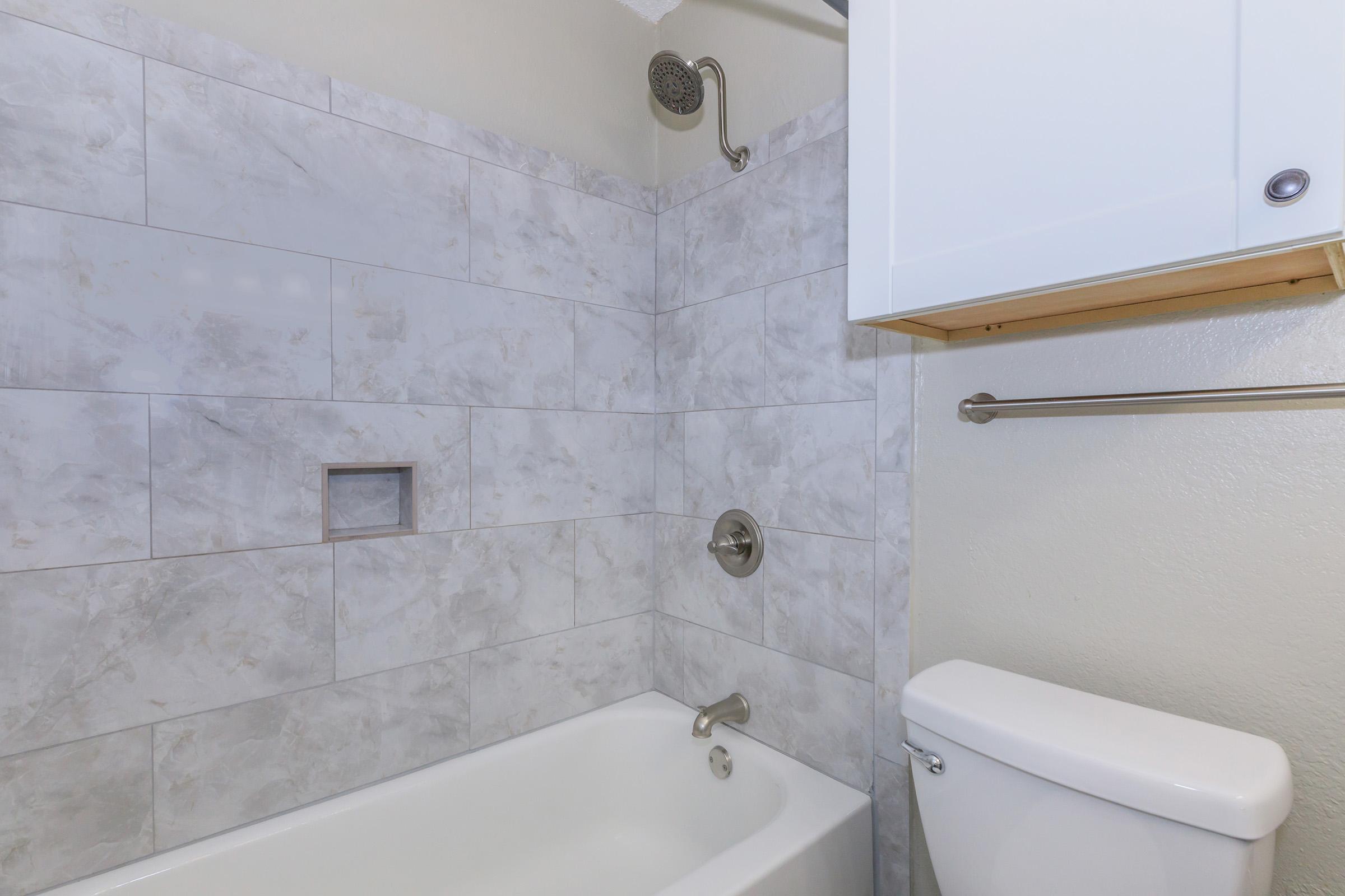 A clean bathroom with a bathtub, a showerhead above, and a toilet nearby. The walls are tiled in a light gray with a marble texture, and there is a small shelf or cabinet above the toilet for storage. Natural light brightens the space.
