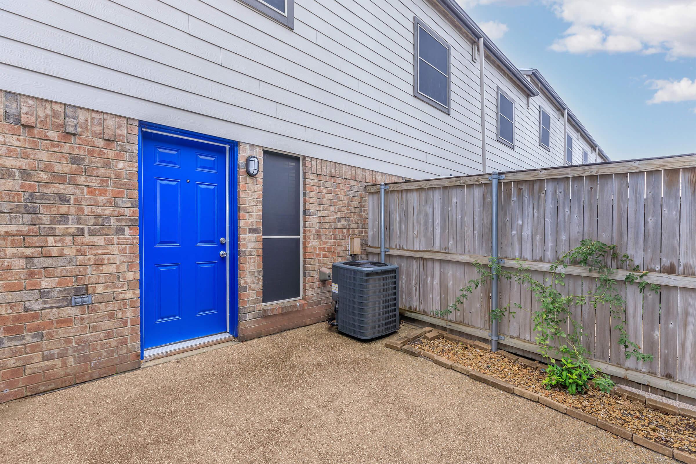 A bright blue door is positioned at the side of a building, made of beige brick and white siding. A plastic trash bin is next to the door, and a wooden fence runs along the side with a small plant climbing on it. The area is well-maintained with gravel and a clean pathway.