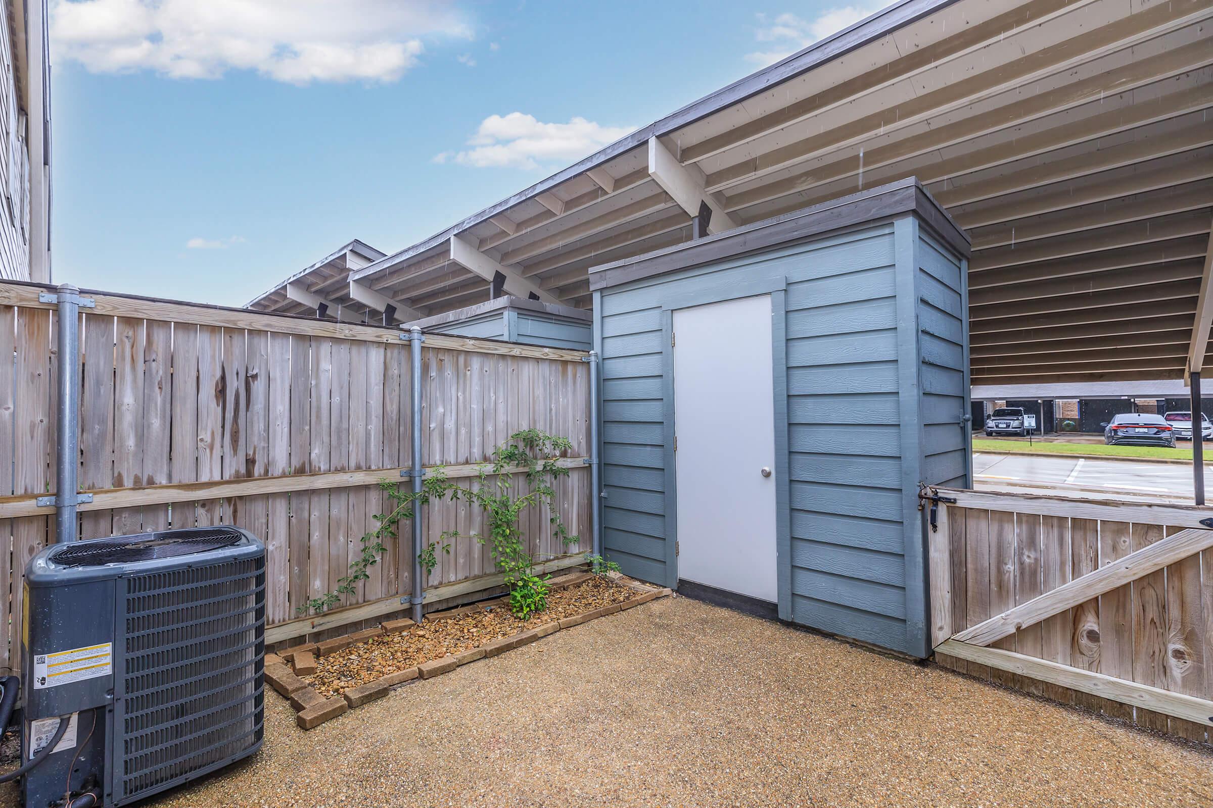 View of a small outdoor space featuring a gray storage shed with a white door, surrounded by a wooden fence. There's a gravel path leading to the shed and a small plant growing nearby. The background shows a structured top with wooden beams. Overall, the area appears clean and well-maintained.