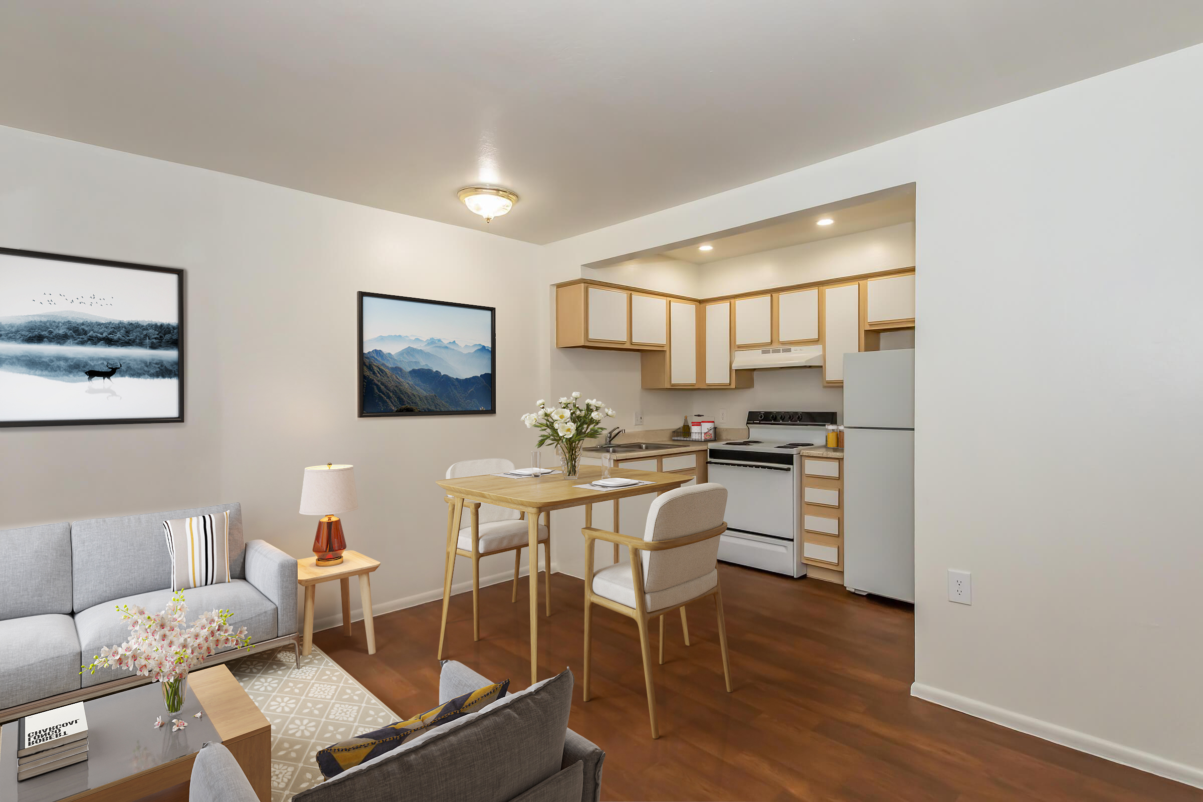 A bright, modern apartment interior featuring a cozy living area with a gray sofa and a small coffee table adorned with flowers. A dining table with two chairs is positioned in front of the kitchen, which includes light wood cabinetry, a white refrigerator, and a stove. Mountain-themed artwork decorates the walls.