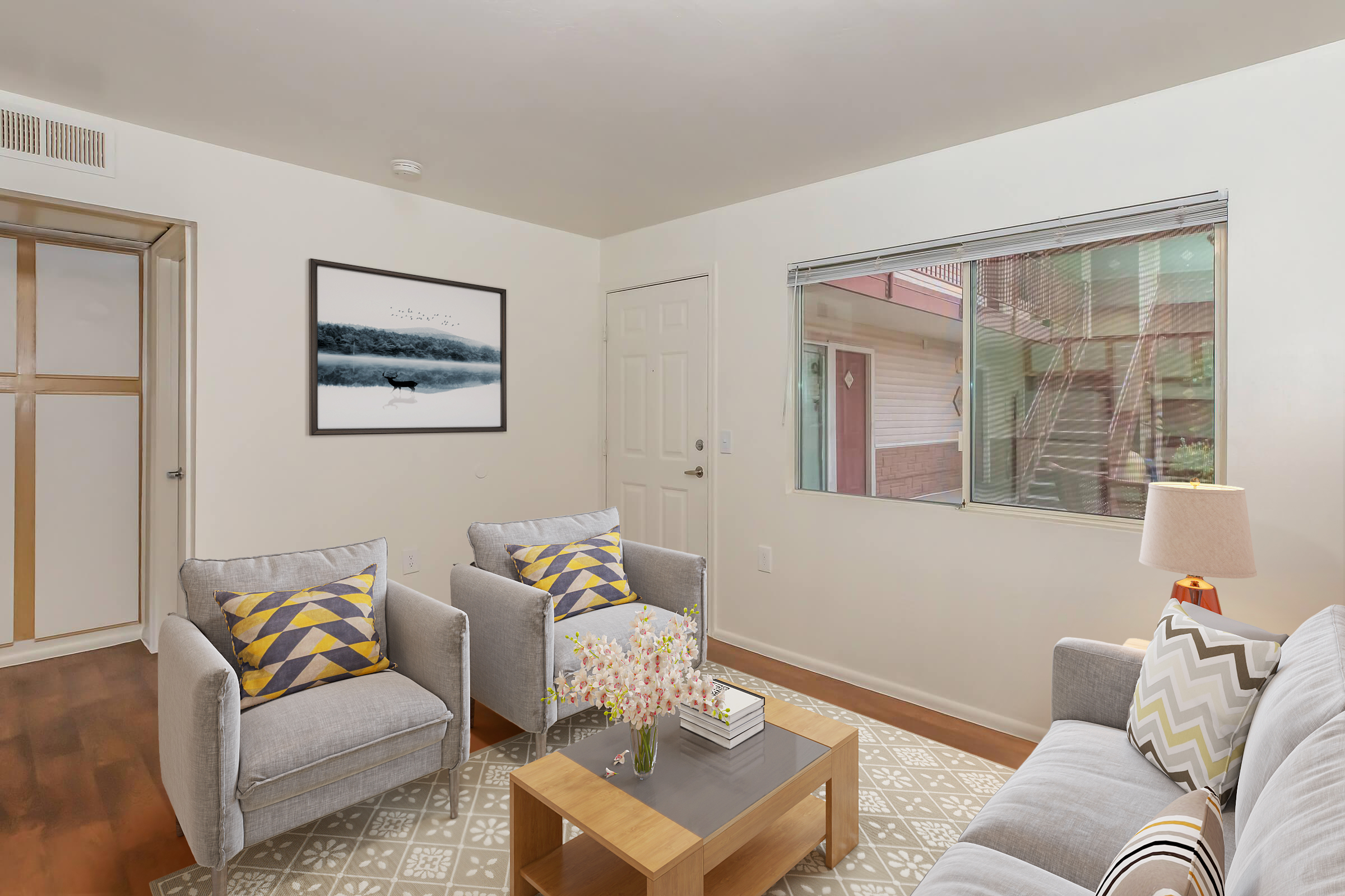 A cozy living room with two gray armchairs and a light gray sofa adorned with yellow and white cushions. A wooden coffee table sits in the center, featuring a vase of flowers and a book. A large framed black-and-white photograph hangs on the wall, and there's a window showing a glimpse of a patio outside.