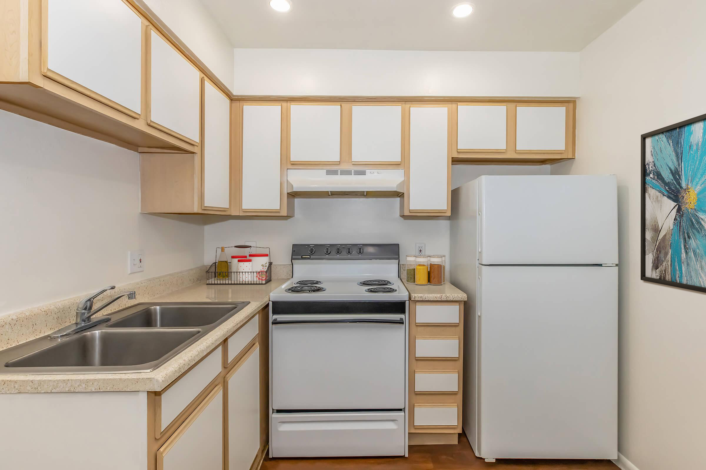 A compact kitchen featuring light wood cabinetry, a double sink, an oven with a stovetop, a refrigerator, and a decorative wall art piece. The countertops are beige, and the overall color scheme is neutral, creating a bright and functional space.