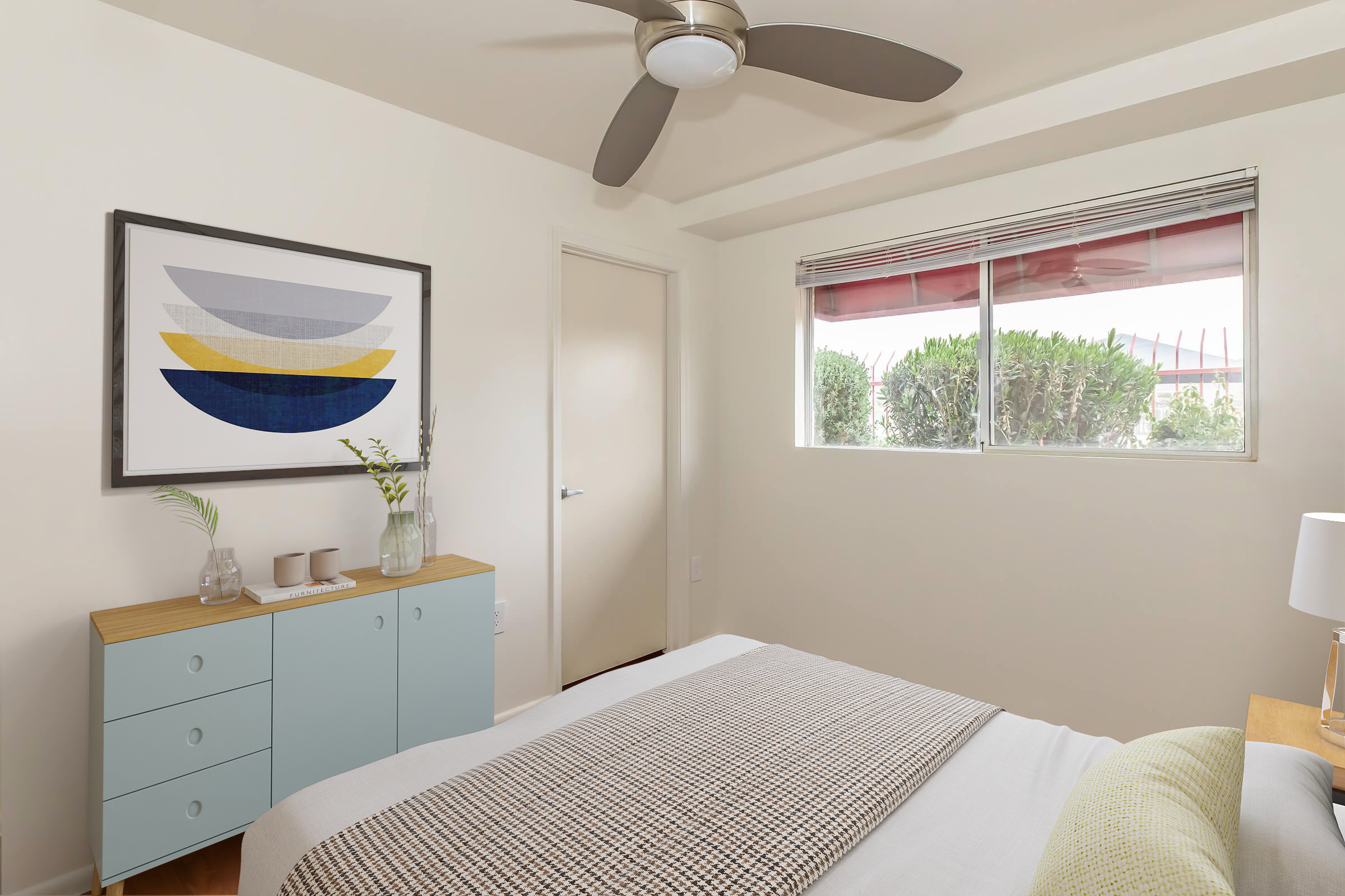 A modern bedroom featuring a neatly made bed with a textured blanket, a light-colored nightstand with a lamp, and a dresser with a soft blue finish. A large piece of abstract art hangs above the dresser. A window with blinds lets in natural light, revealing greenery outside.