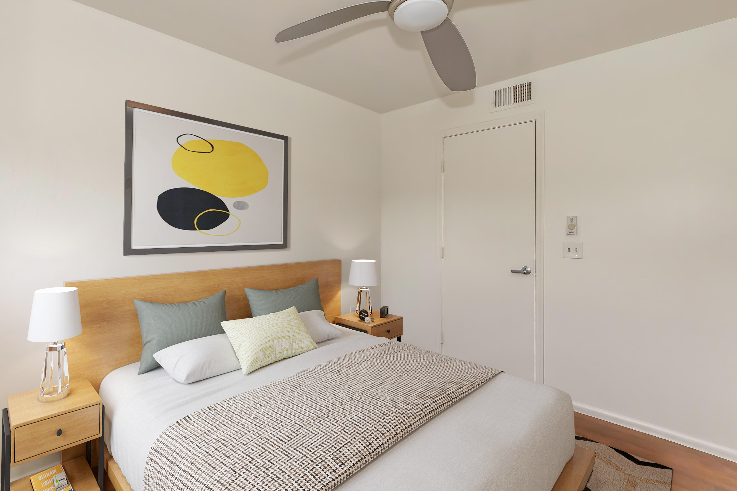 A modern bedroom featuring a neatly made bed with light-colored bedding, two bedside tables with lamps, and an abstract wall art featuring yellow and black shapes. A ceiling fan is visible, and the room has neutral-colored walls and wood flooring.