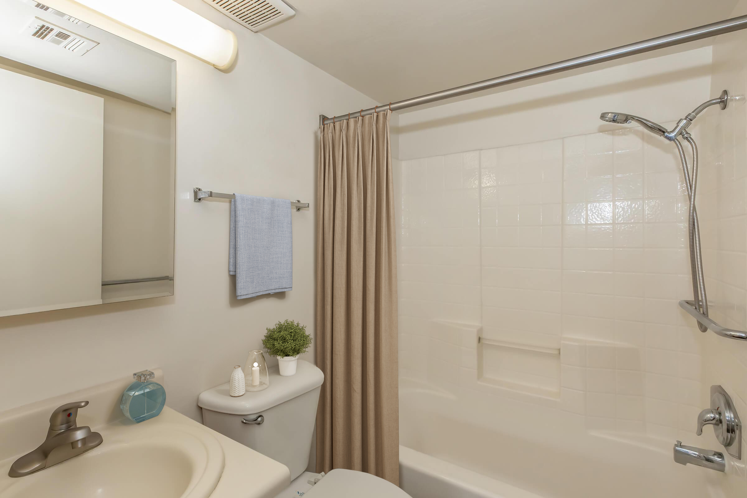 A clean and modern bathroom featuring a bathtub with a shower, a toilet, a sink with a faucet, and a mirror. There is a light-colored shower curtain, a blue hand towel on a rack, a small plant, and a few decorative toiletries on the sink. The walls are painted white, creating a bright atmosphere.
