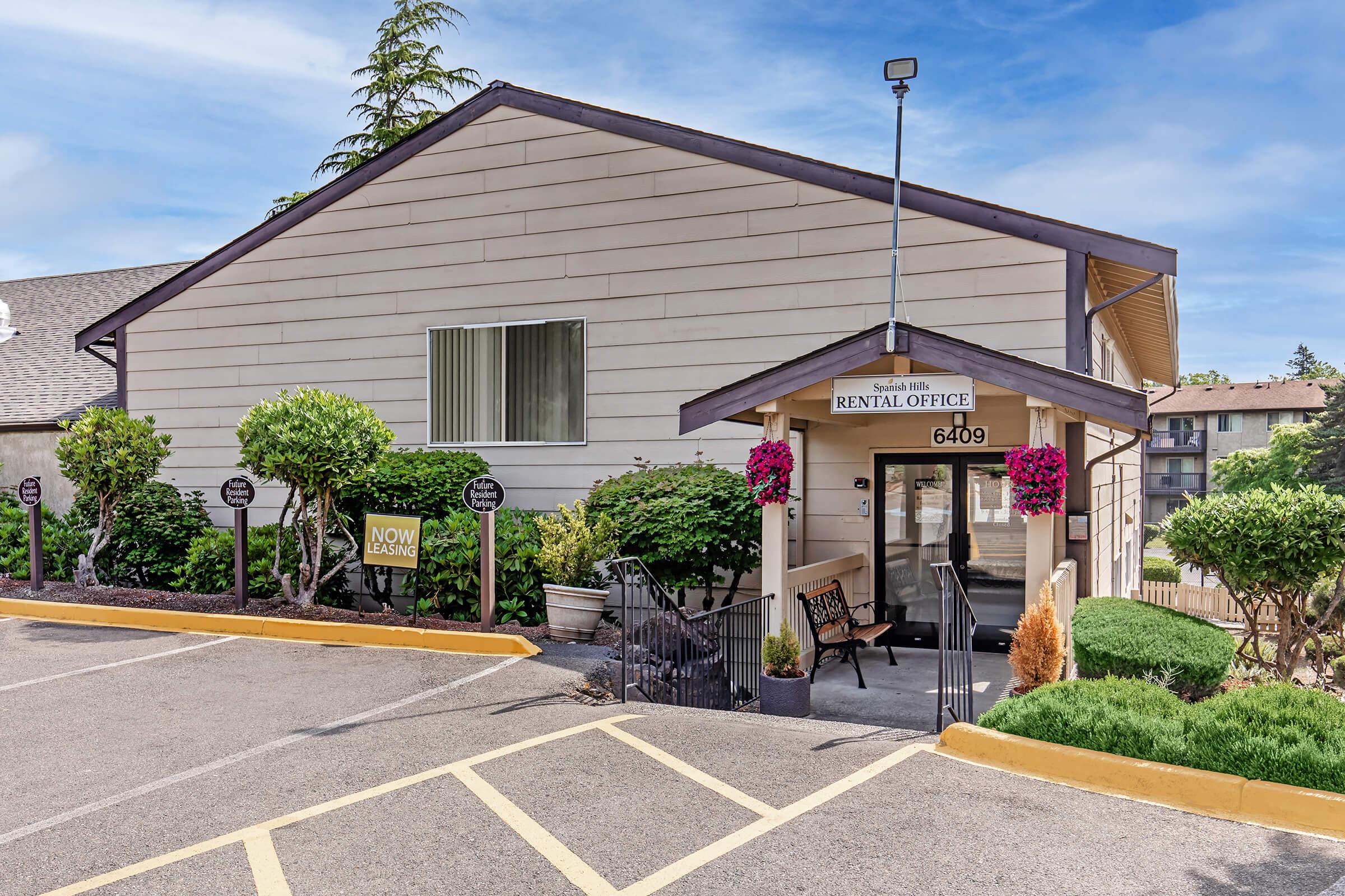 Exterior view of a rental office building. The structure is a single-story with light tan siding, featuring a sign that reads "RENTAL OFFICE." Flower beds and bushes frame the entrance, which has a small staircase. Parking spaces are visible in front, marked with “NOW LEASING” signage.