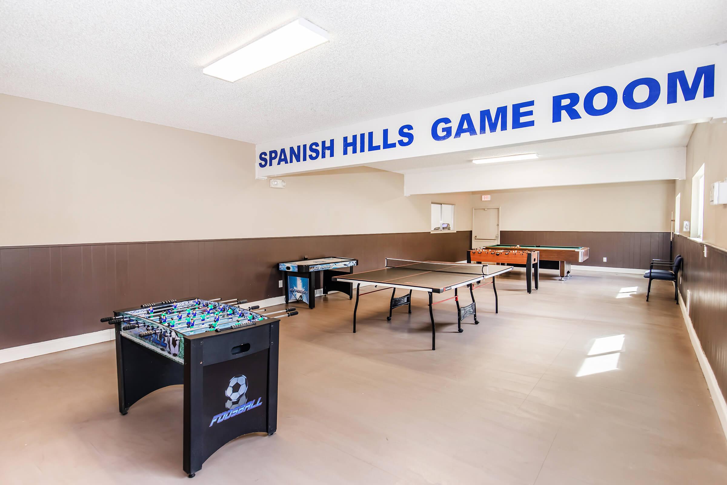 An interior view of the Spanish Hills Game Room, featuring a foosball table, a ping pong table, and an air hockey table. The space is well-lit with simple decor and a sign indicating the room's name. The walls are painted in neutral tones, and the floor is light-colored, providing a clean and welcoming atmosphere for recreational activities.