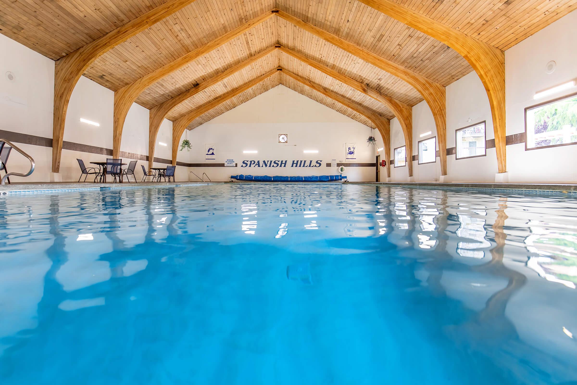 Indoor swimming pool at Spanish Hills featuring a wooden beam ceiling. The clear blue water reflects the light, with chairs arranged on the side. A pool cover is visible at the far end, and a clock is mounted on the wall, creating a relaxing atmosphere.
