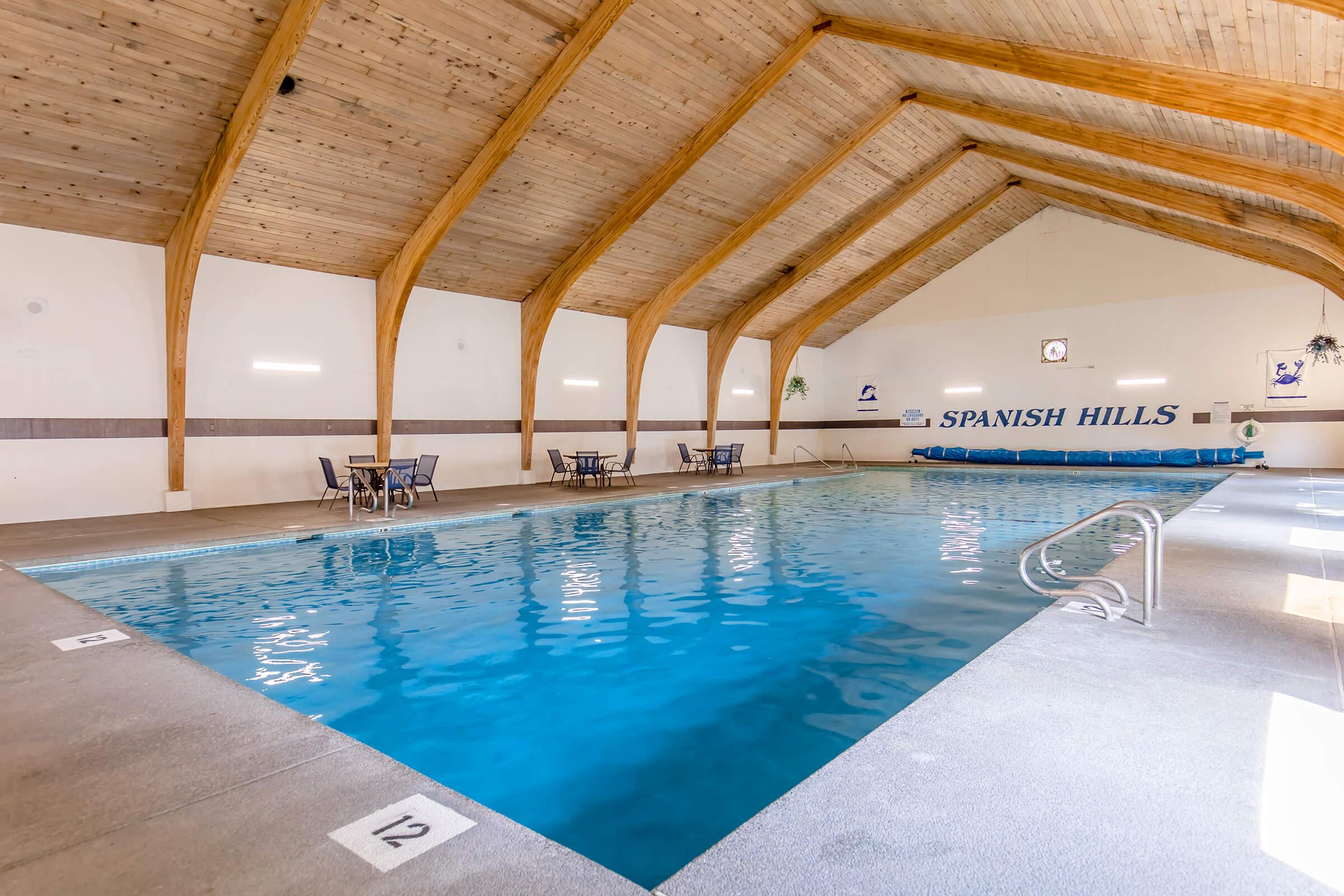 Indoor swimming pool at Spanish Hills, featuring a wooden ceiling with exposed beams. The pool area includes lounge chairs and tables, with a clear blue water inviting for swimming. The walls are decorated minimally, enhancing the spacious and bright atmosphere of the facility.