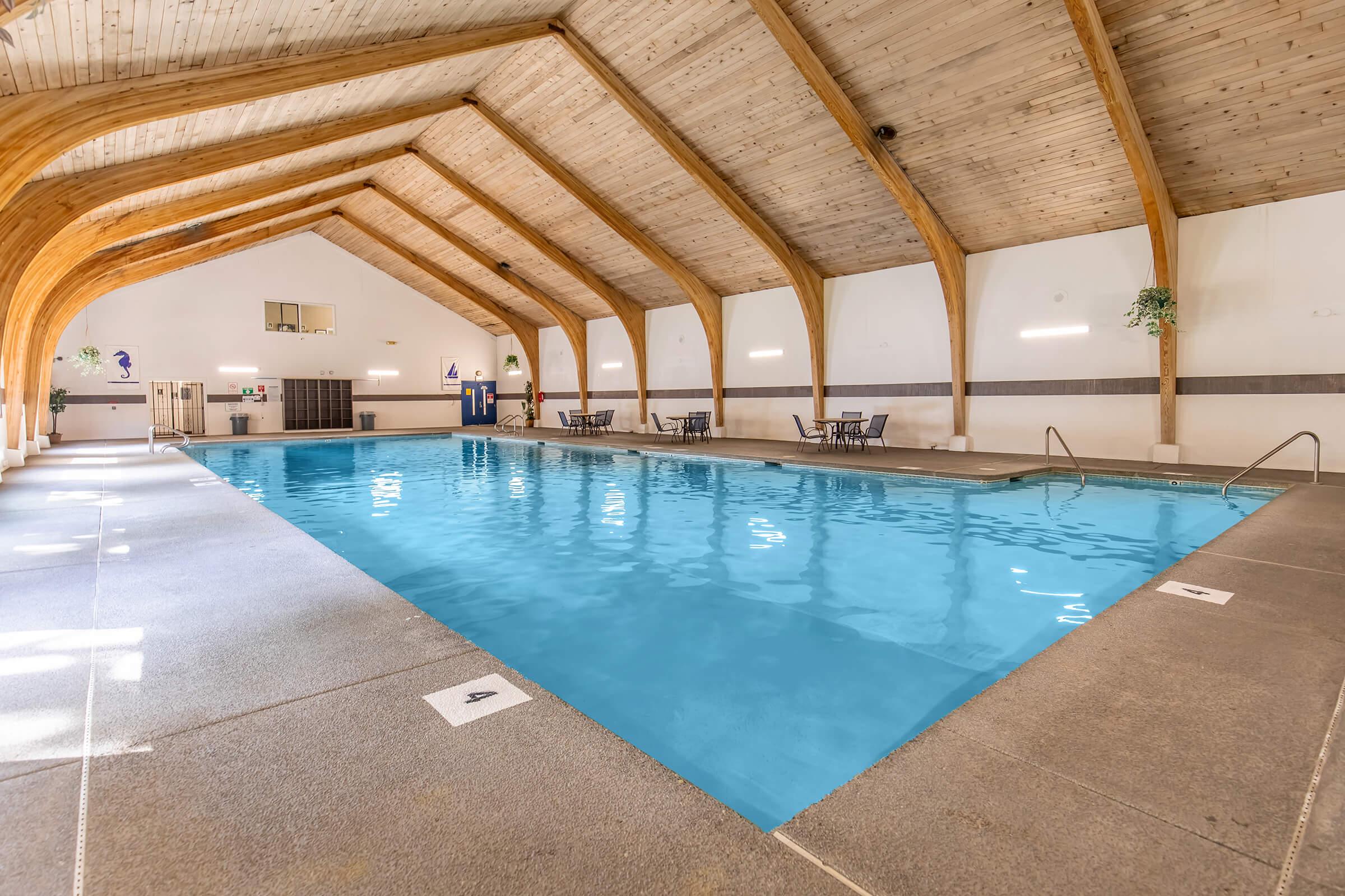 Indoor swimming pool with clear blue water, surrounded by a wooden framework and high ceilings. Sunlight streams in, creating reflections on the water's surface. There are chairs and tables along the poolside, offering a relaxing atmosphere for guests.
