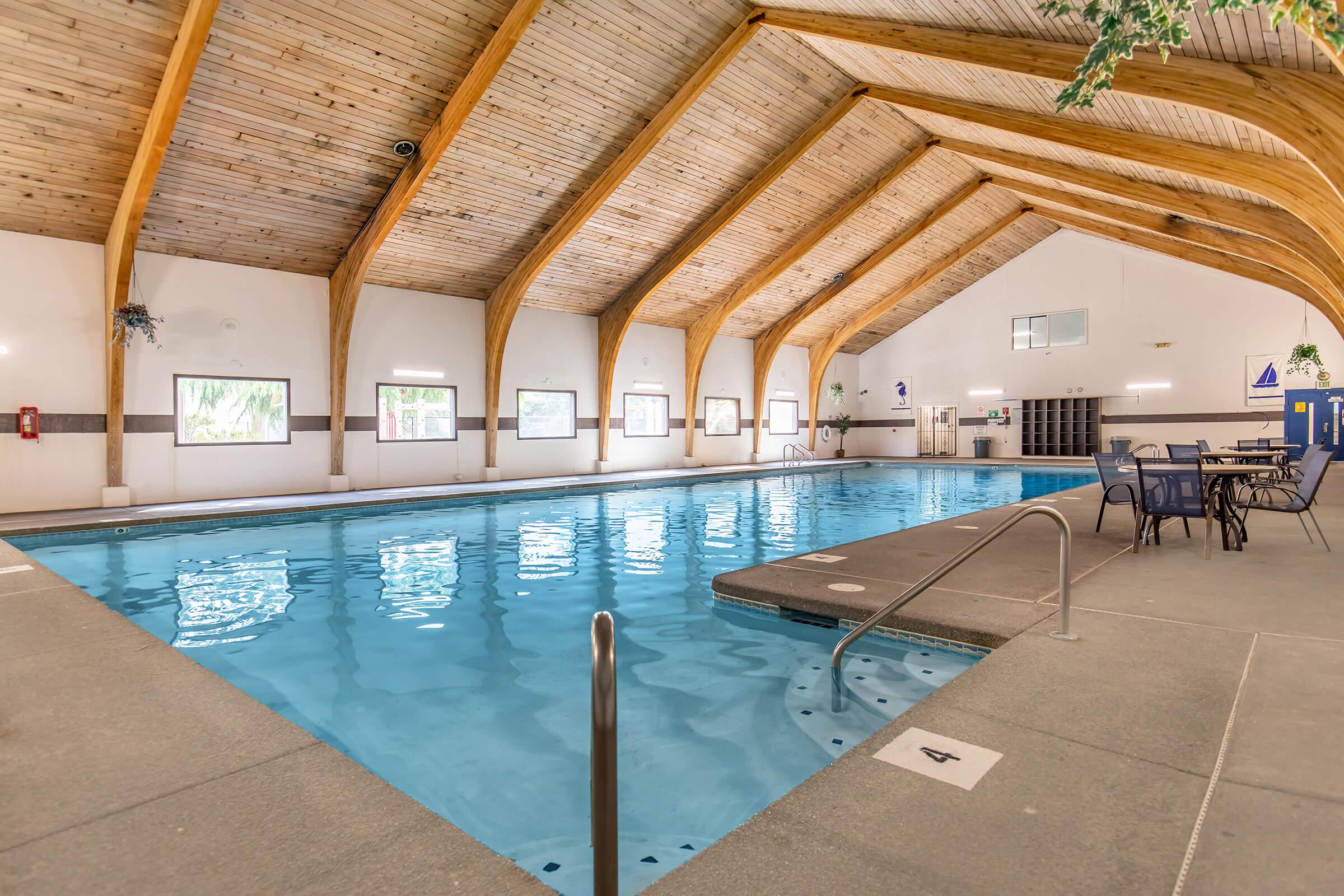 Indoor swimming pool with clear blue water, surrounded by a stone deck. The ceiling features wooden beams, creating a spacious, airy atmosphere. There are several windows allowing natural light in, and a few lounge chairs are set up nearby. The area is clean and well-maintained, inviting relaxation and recreation.