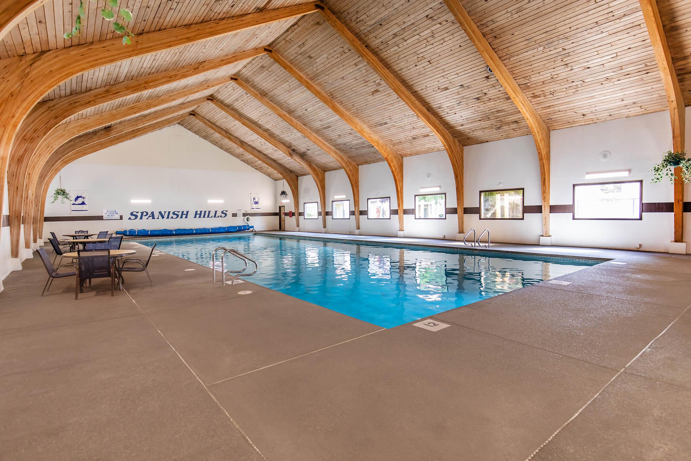 Indoor swimming pool at Spanish Hills, featuring wooden beams and a smooth concrete floor. There are tables and chairs near the pool area, with large windows letting in natural light. The water is clear, and the atmosphere is inviting and well-maintained.