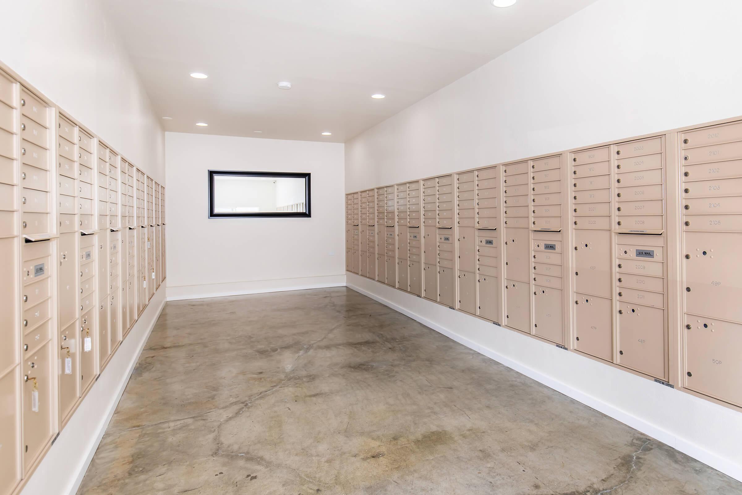 A hallway featuring multiple beige mailboxes on either side, with a large mirror at the far end, illuminated by overhead lights. The floor is concrete, and the walls are painted white, creating a clean and organized space.