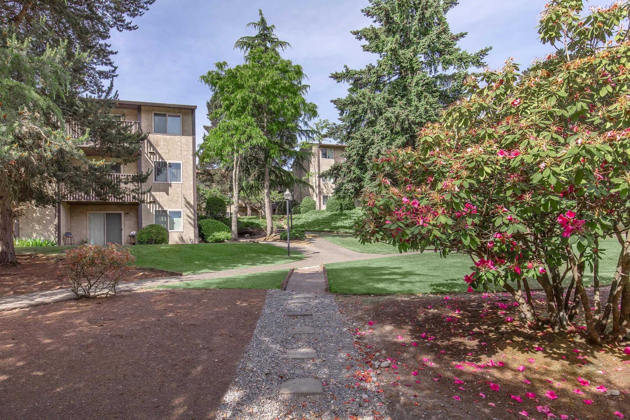 A landscaped pathway leads through a lush green area with flower beds, shrubs, and trees. In the background, there are two apartment buildings surrounded by well-maintained lawns and vibrant pink and purple flowers, creating a pleasant residential atmosphere.