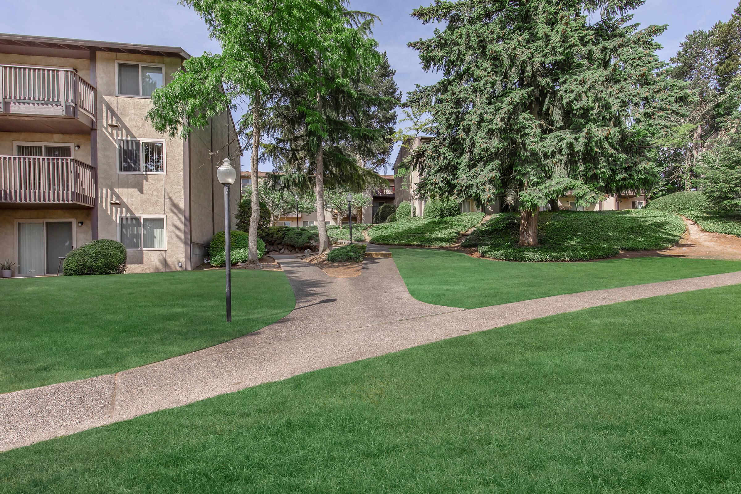 A landscaped area featuring two apartment buildings surrounded by lush green lawns and trees. A paved pathway winds through the grass, connecting the buildings, with a lamppost visible along the path. The scene is bright and inviting, showcasing a residential community atmosphere.