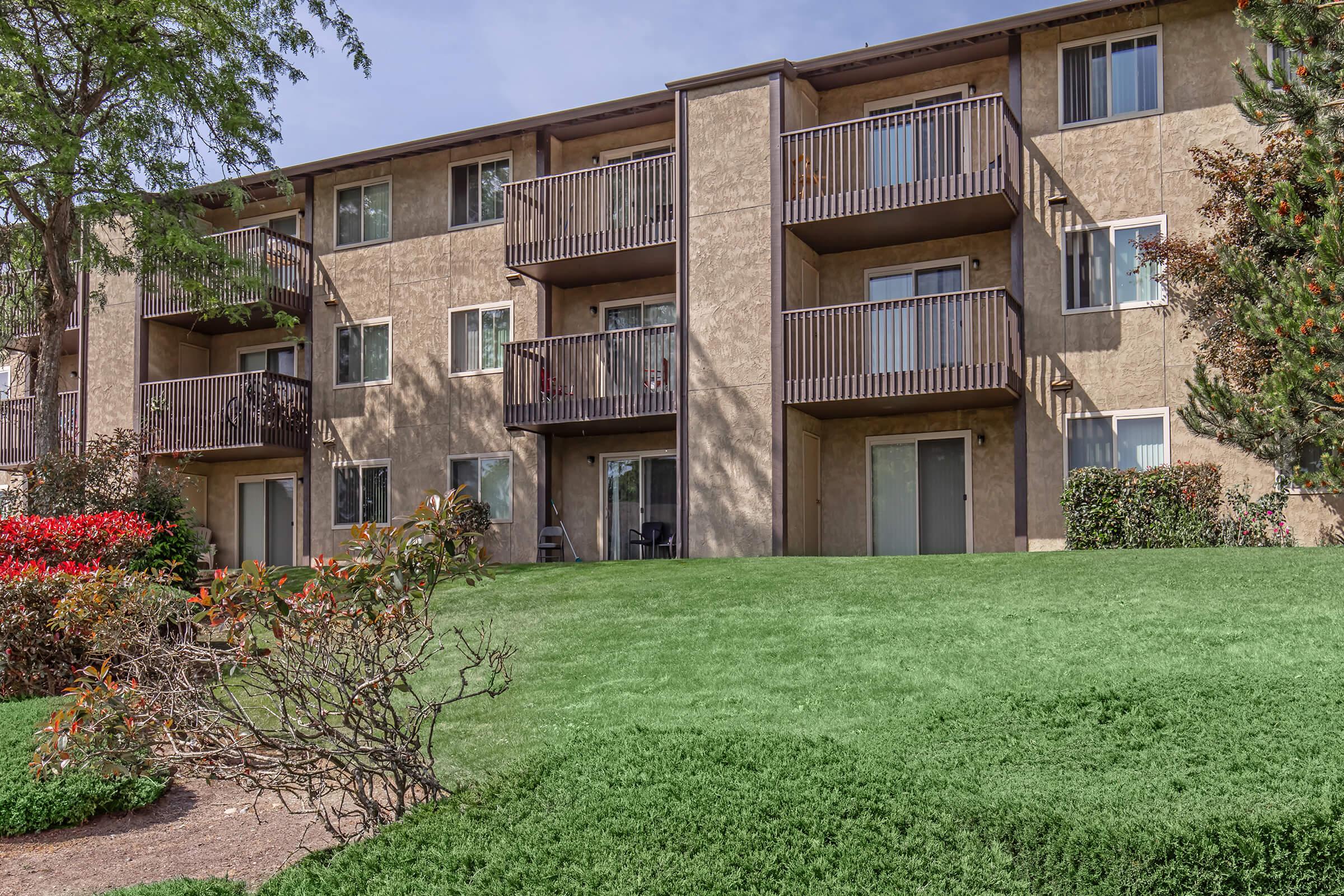 A multi-story residential building with balconies and large windows, set in a landscaped area featuring green grass and flowering shrubs. The building has a warm, neutral exterior color and is surrounded by trees and bushes, creating a welcoming environment.