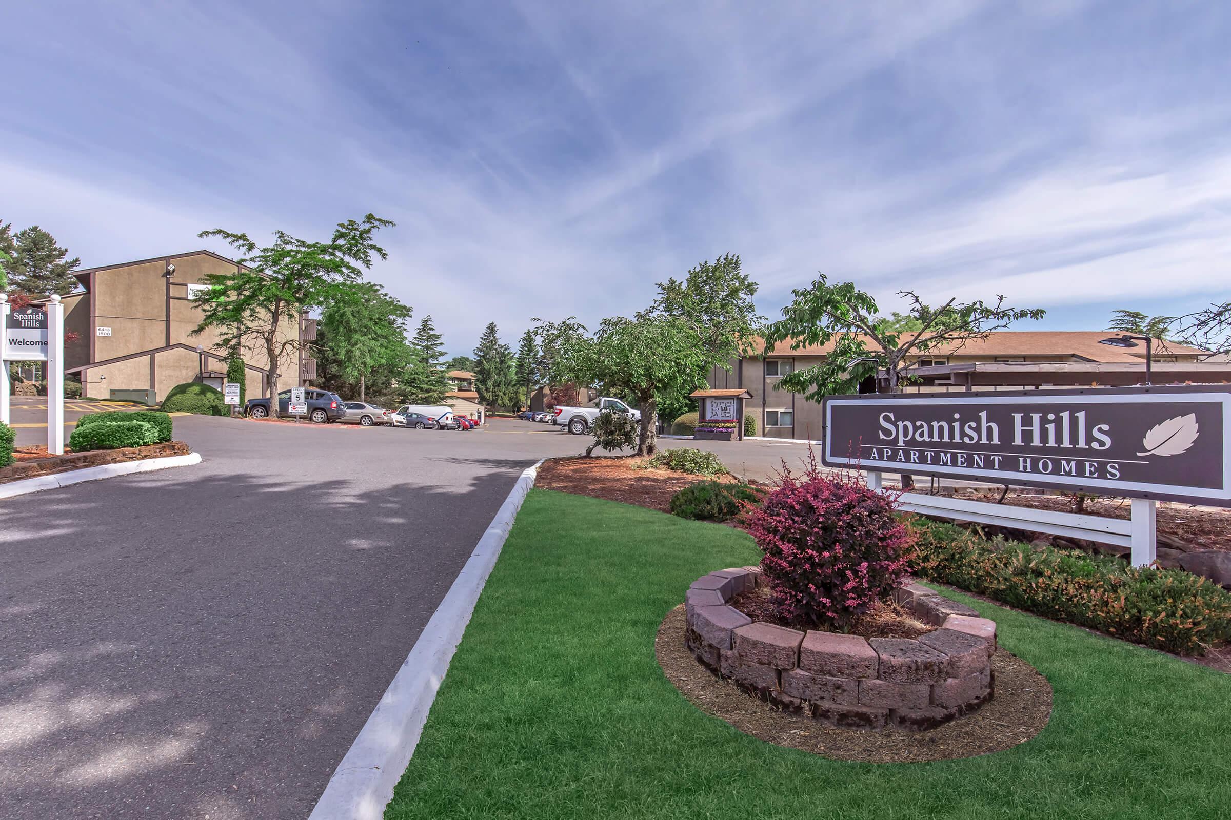 A view of the entrance to Spanish Hills Apartment Homes, featuring a welcoming sign. The image shows well-maintained landscaping, a paved road leading into the complex, and trees lining the pathway. Apartments are visible in the background, set against a clear blue sky.