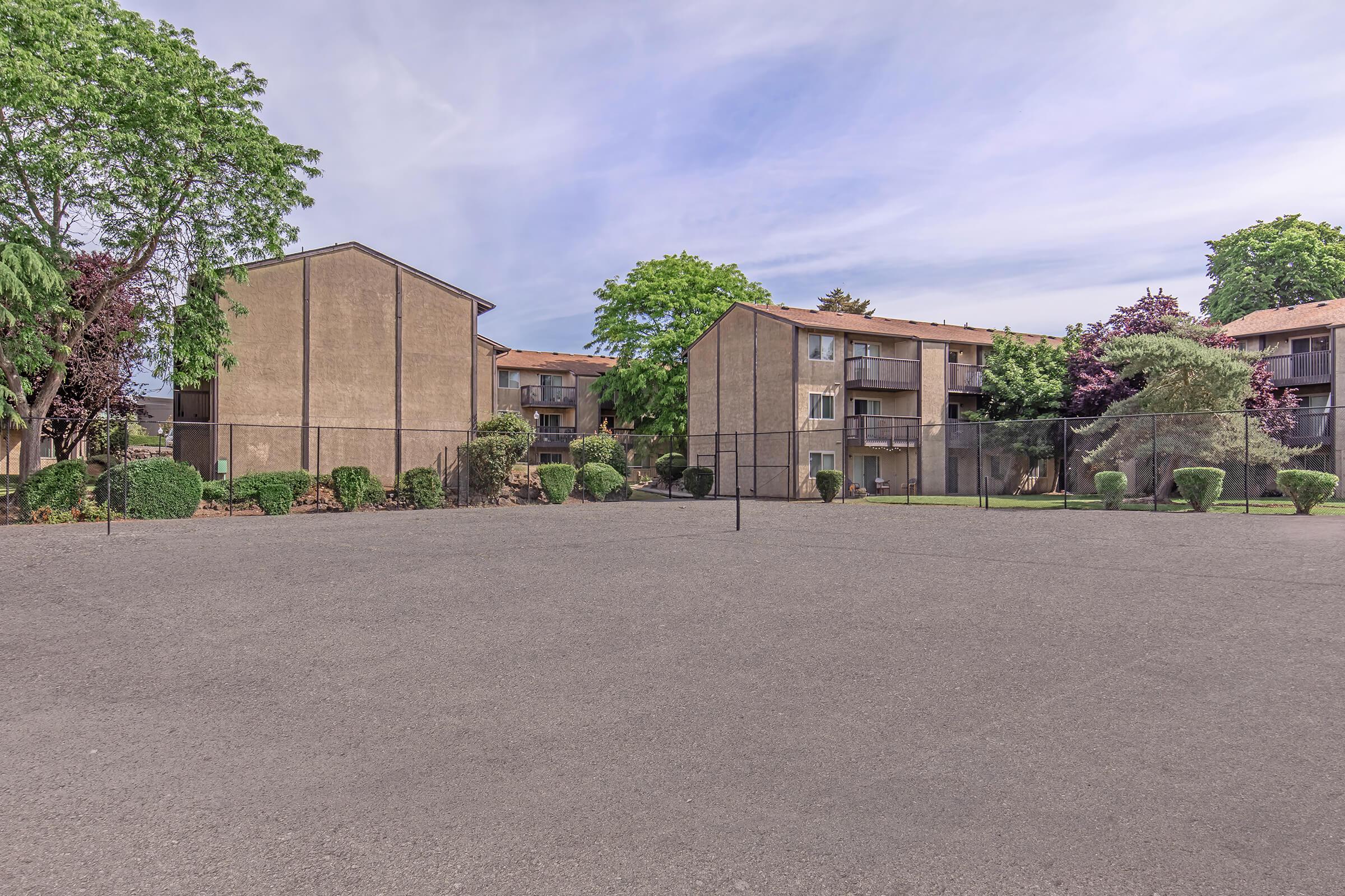 View of an empty outdoor tennis court surrounded by two apartment buildings and well-maintained greenery, with trees and shrubs lining the area. The sky is partly cloudy, creating a serene atmosphere in the residential setting.
