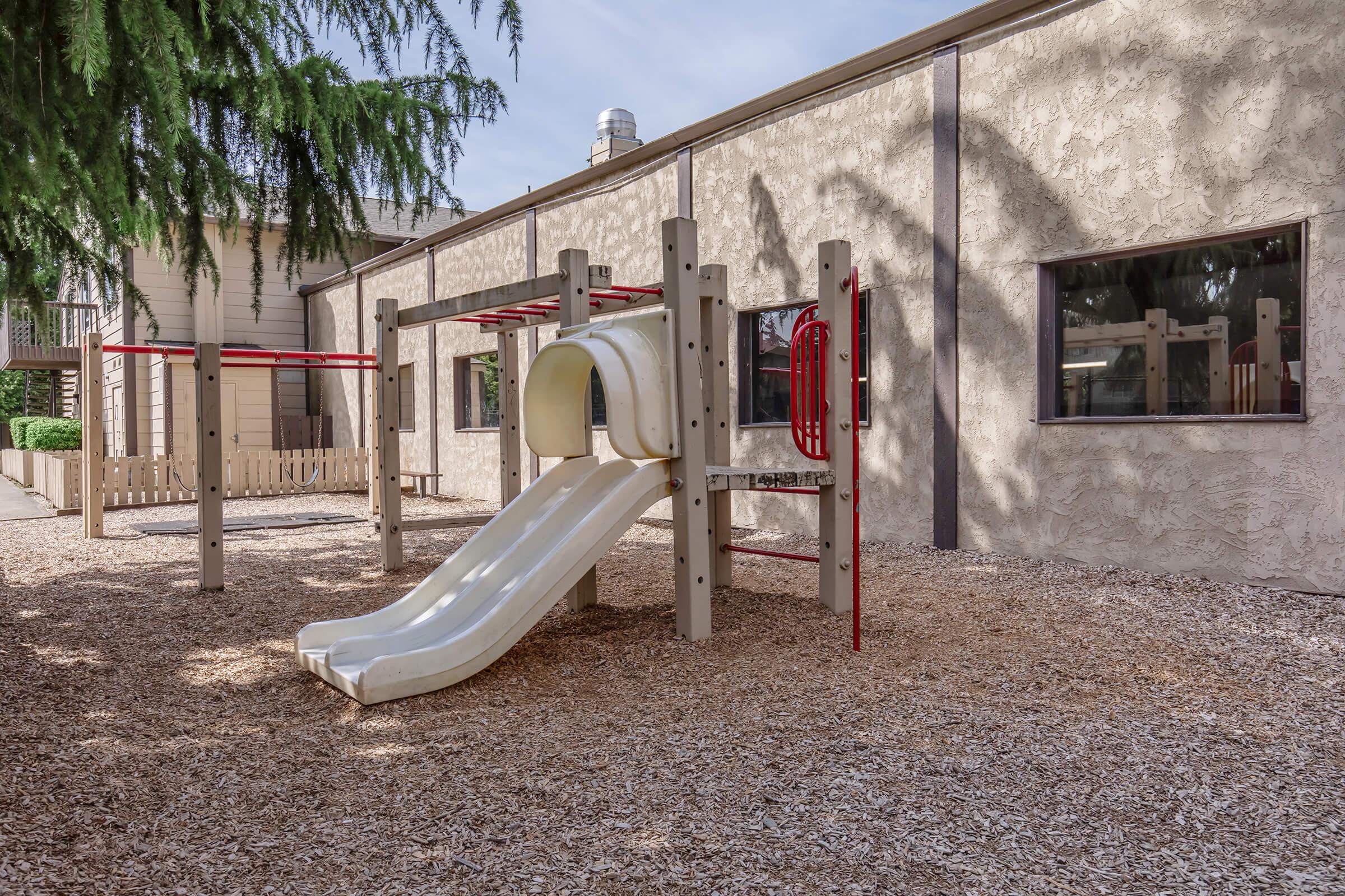 Playground equipment featuring a white slide and climbing structure, set on a mulch-covered ground. In the background, there is a textured wall of a building with large windows and a wooden fence. Some trees provide shade over the play area.