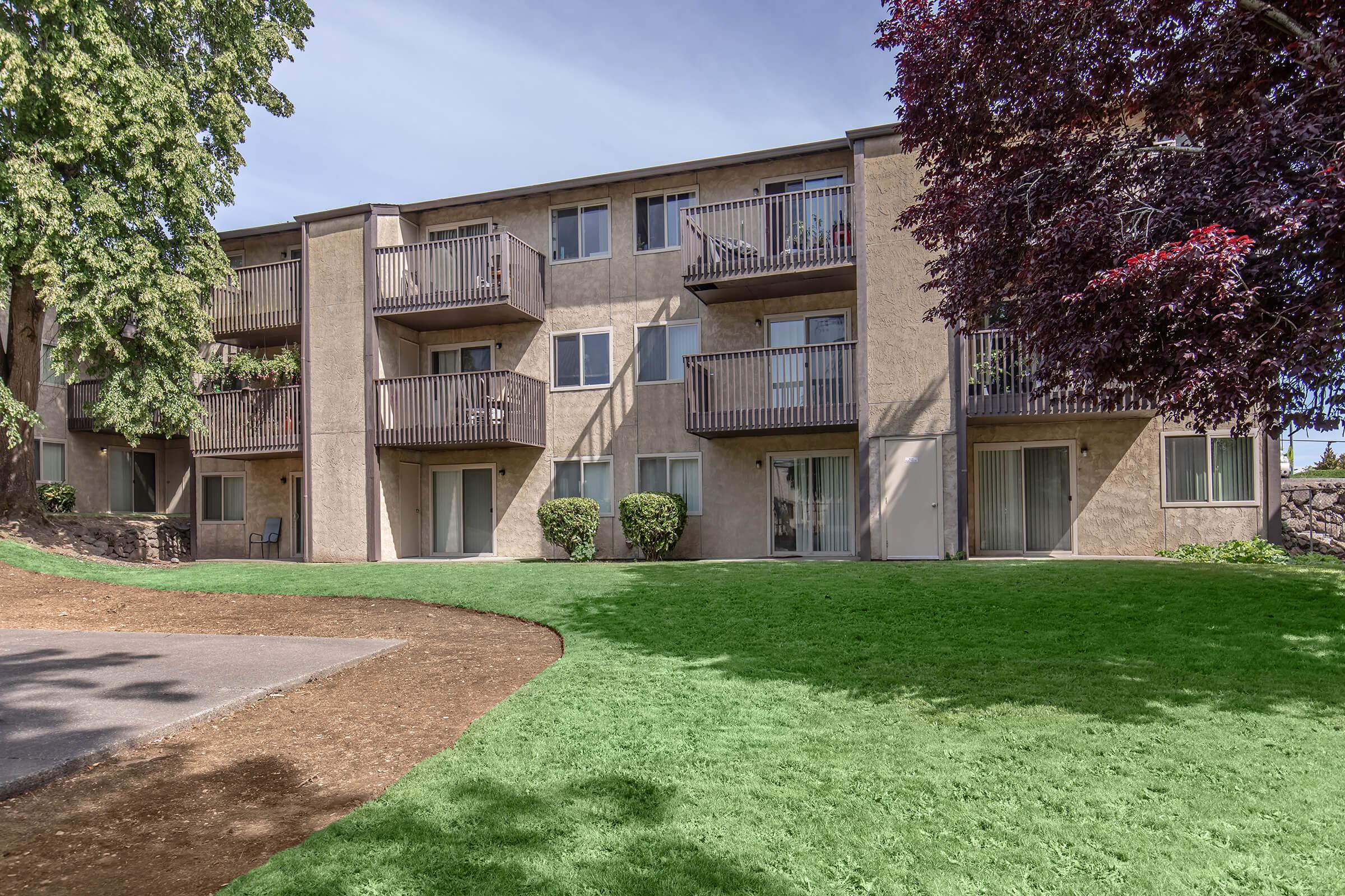 A multi-unit apartment building with beige walls, multiple balconies, and large windows, surrounded by a well-maintained green lawn and trees. The pathway in front is made of gravel, leading to the building entrance.