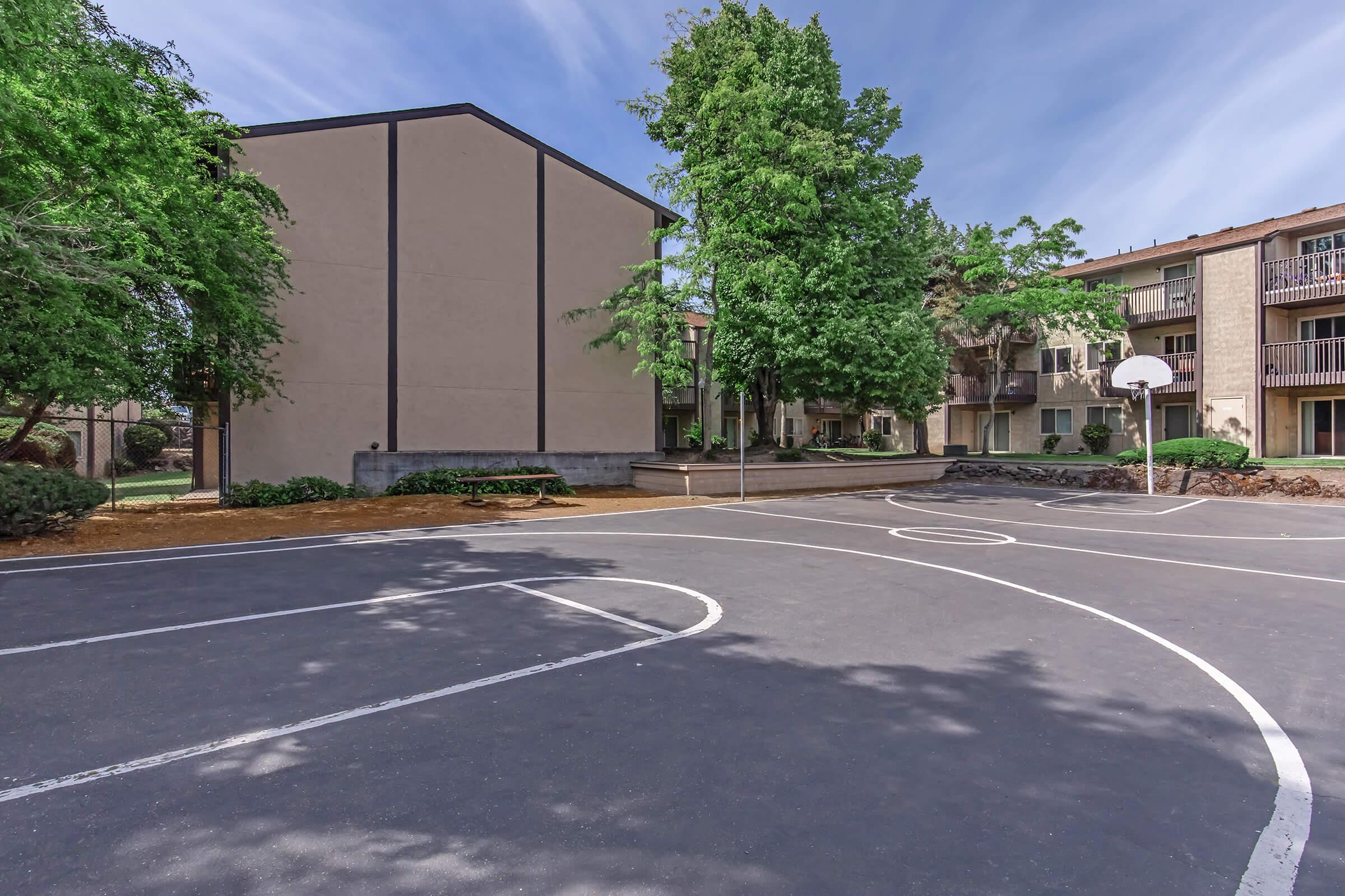 A basketball court surrounded by residential buildings, featuring a single hoop and marked court lines. Lush green trees provide shade, and the sky above is clear with a few clouds. The setting is quiet and inviting, ideal for outdoor activities.