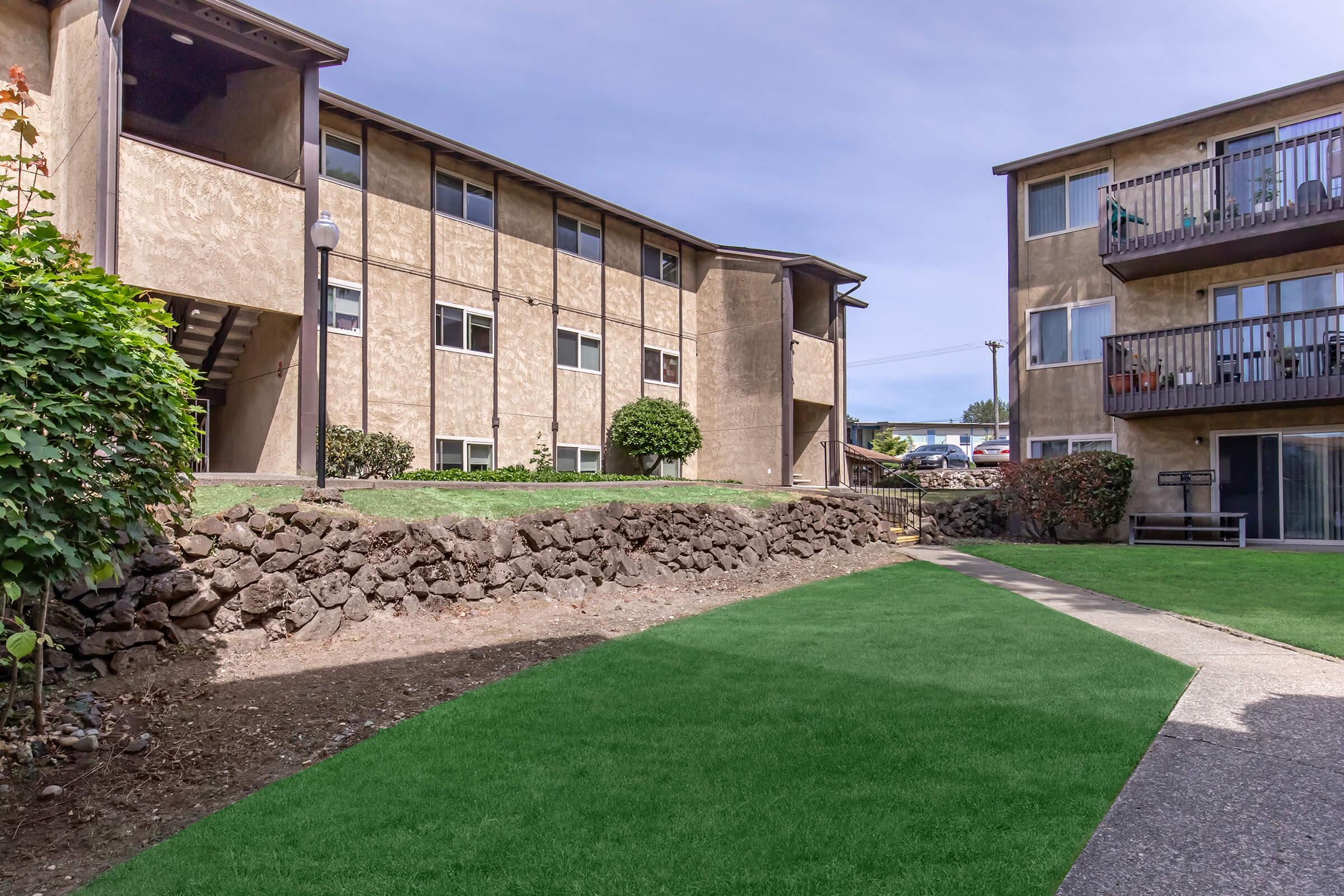 View of a well-maintained apartment complex featuring two buildings with balconies. The scene includes a green lawn area and a path leading through the courtyard, surrounded by rock landscaping and shrubs. Bright day with a clear sky and cars parked in the background.