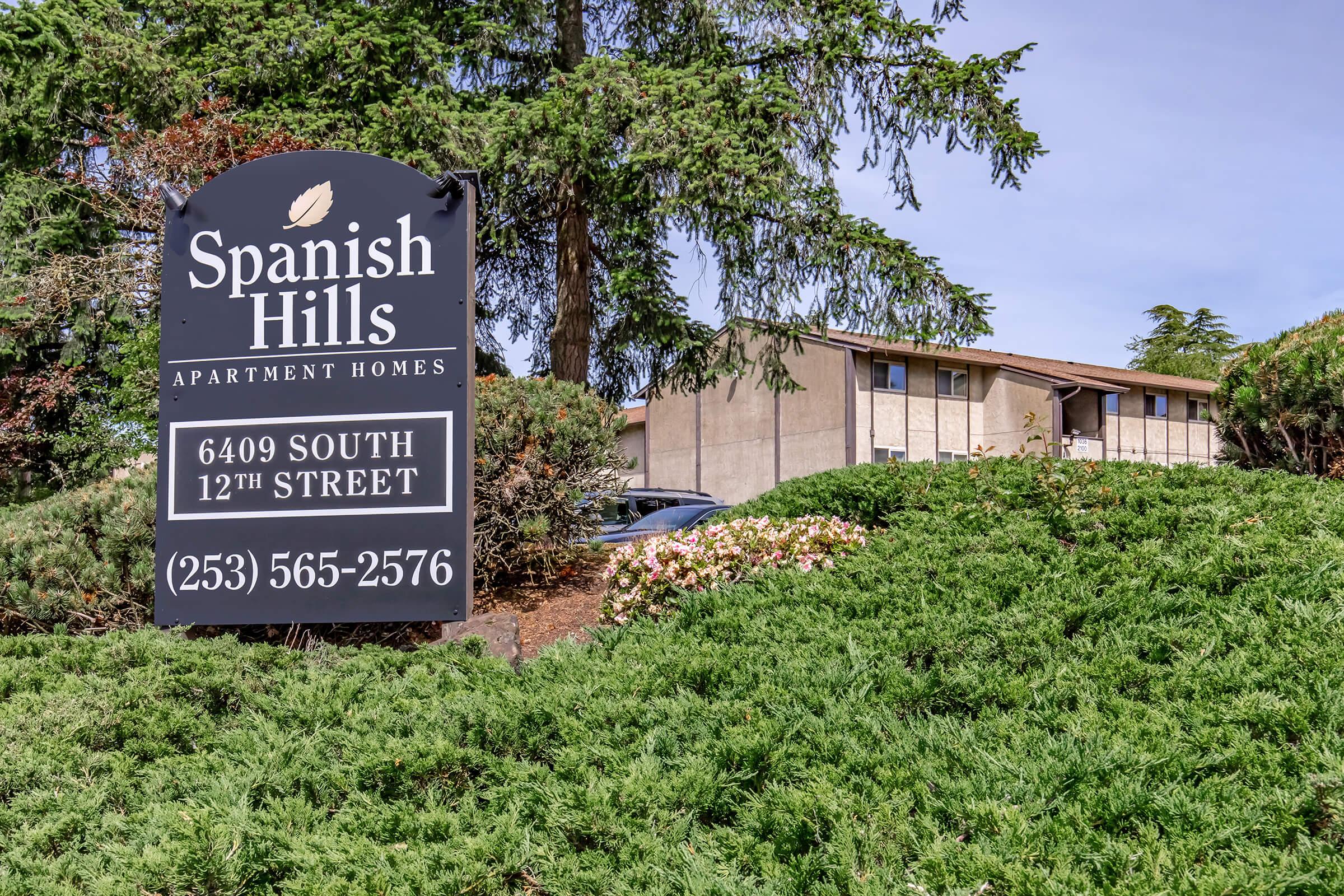 Sign for Spanish Hills Apartment Homes located at 6409 South 12th Street, featuring a dark background with white text. The sign displays the phone number (253) 565-2576. Surrounding the sign are green bushes and trees, with a glimpse of the apartment building in the background.