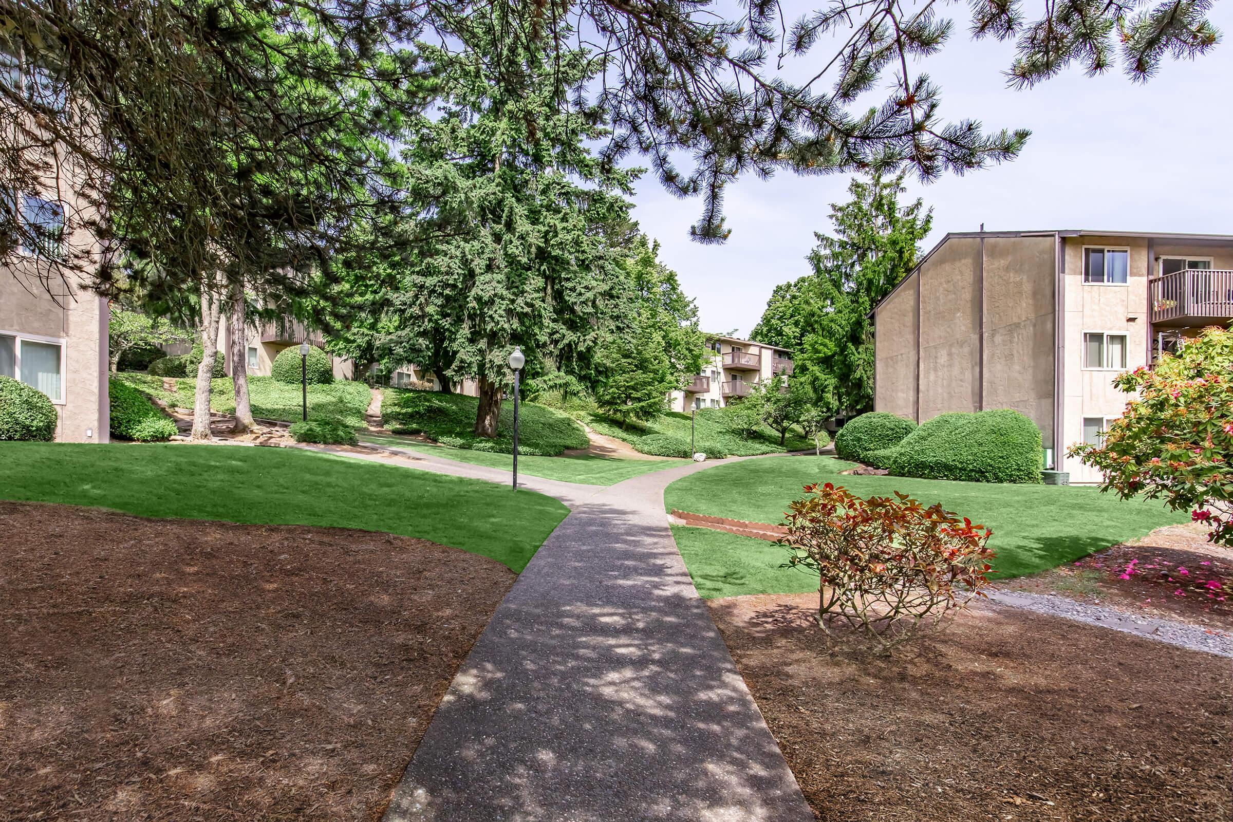 A landscaped pathway winding through a green area, flanked by well-maintained shrubs and trees. On either side, there are two buildings with balconies, suggesting a residential or apartment complex. The sky is clear, and the scene is serene, reflecting a well-kept outdoor space.